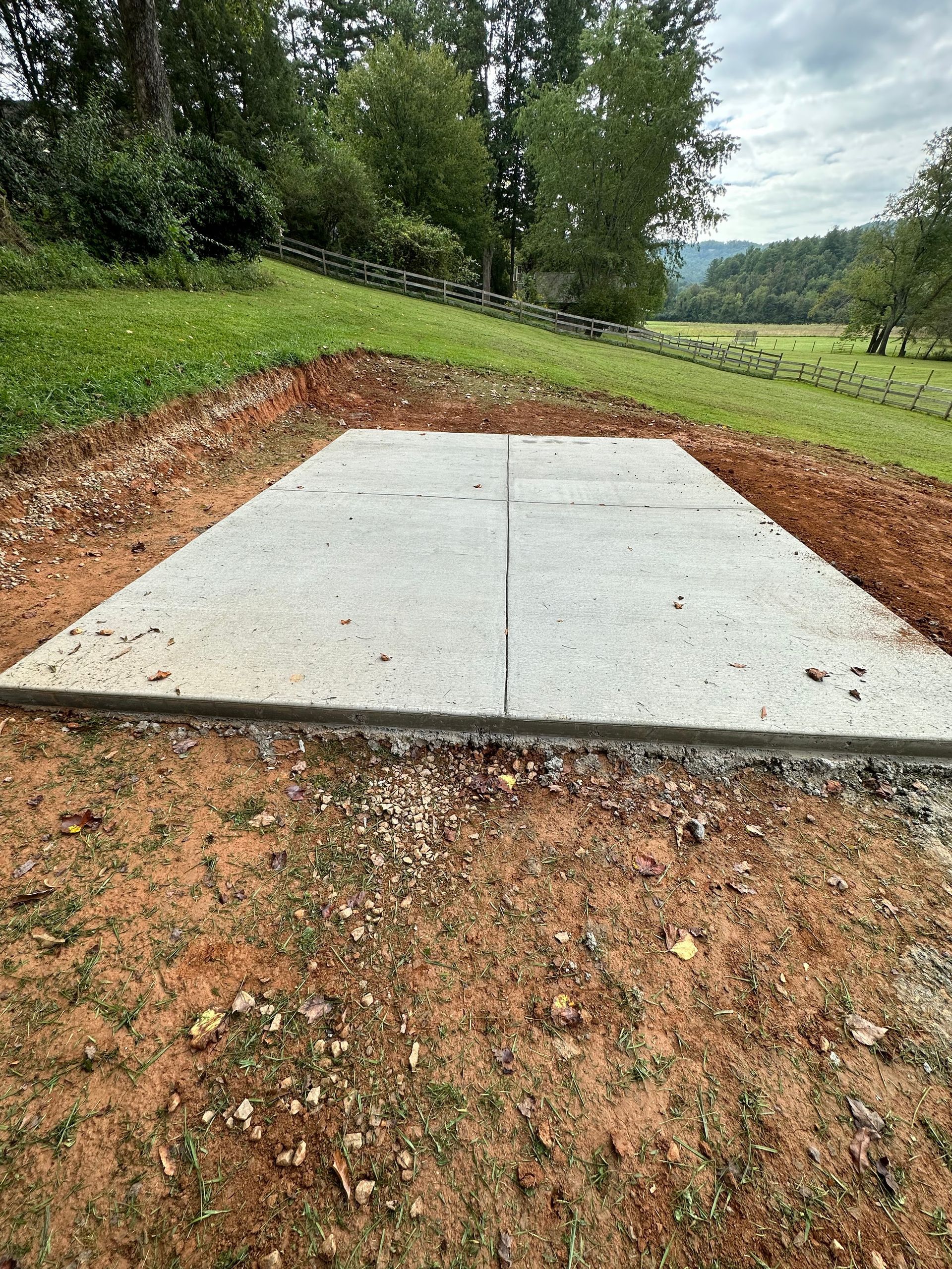 A flat, rectangular concrete slab poured into a grassy hillside, surrounded by exposed dirt and a distant tree line.