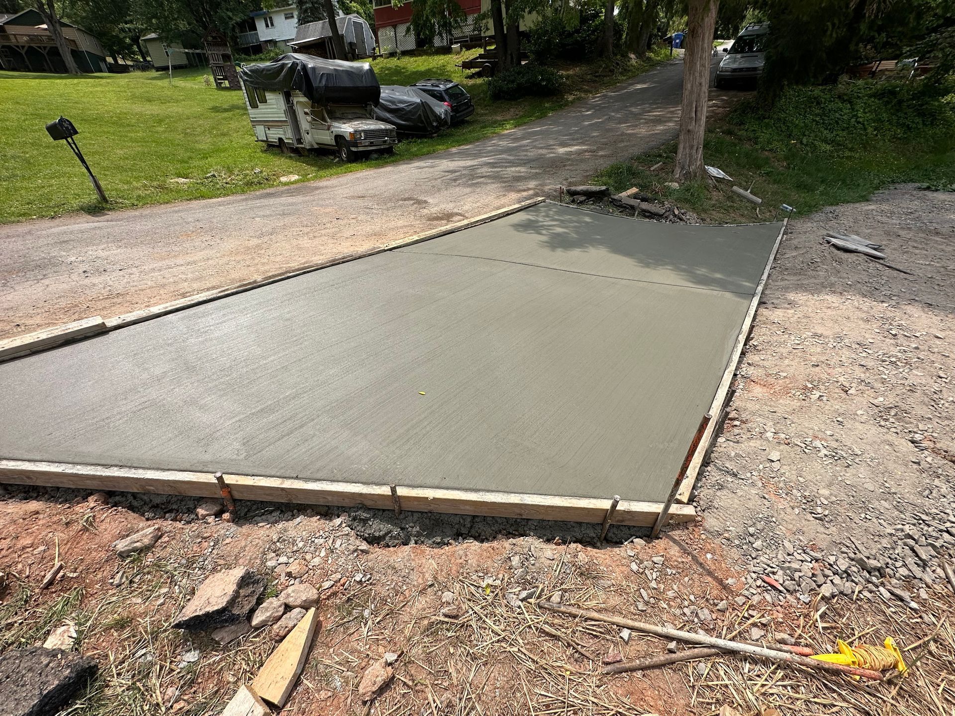 Freshly poured concrete driveway slab inside wooden formwork, located in a gravel area with grass and trees nearby.