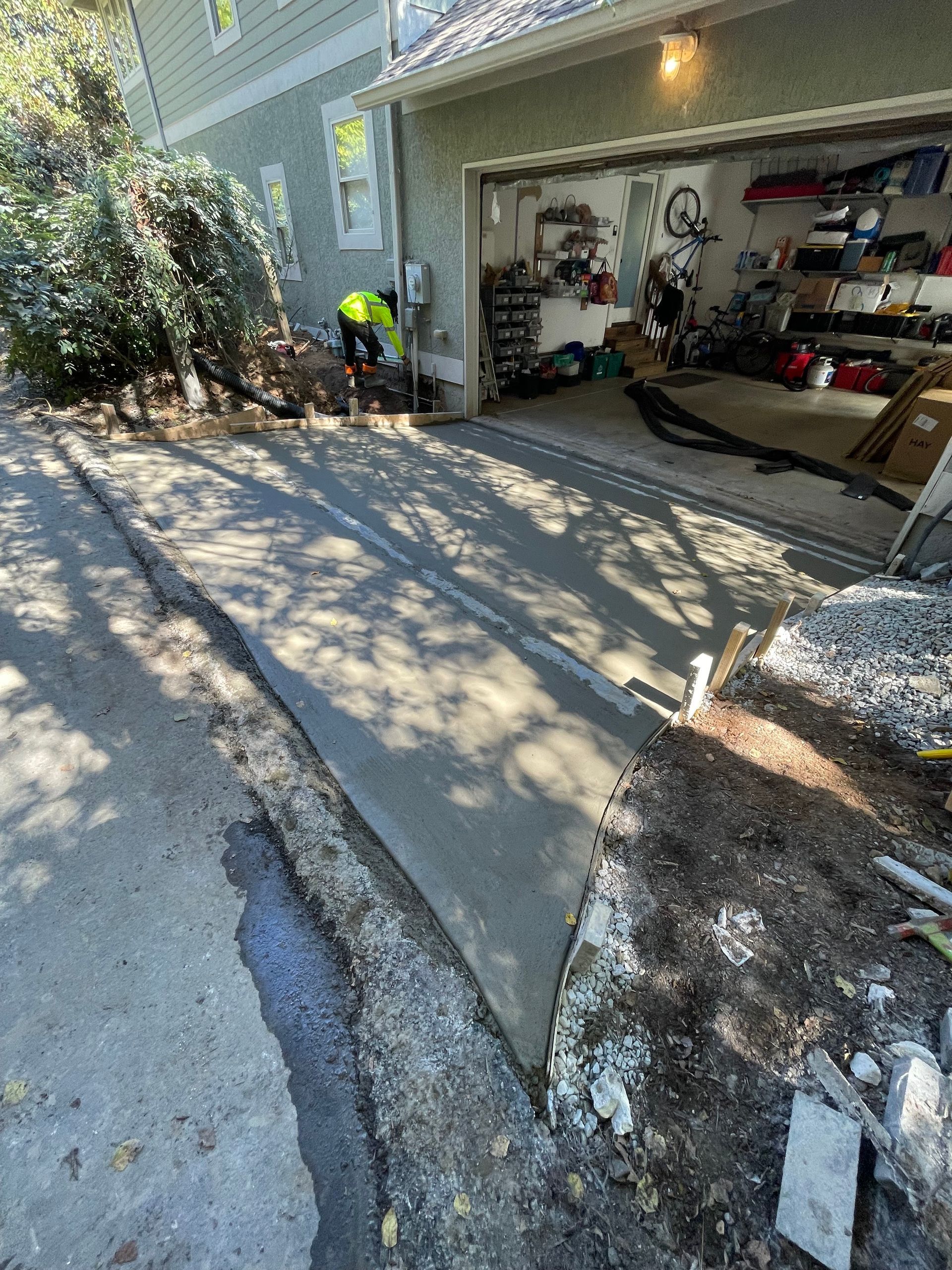 A worker in a yellow safety vest smooths freshly poured concrete on a driveway leading into an open garage.