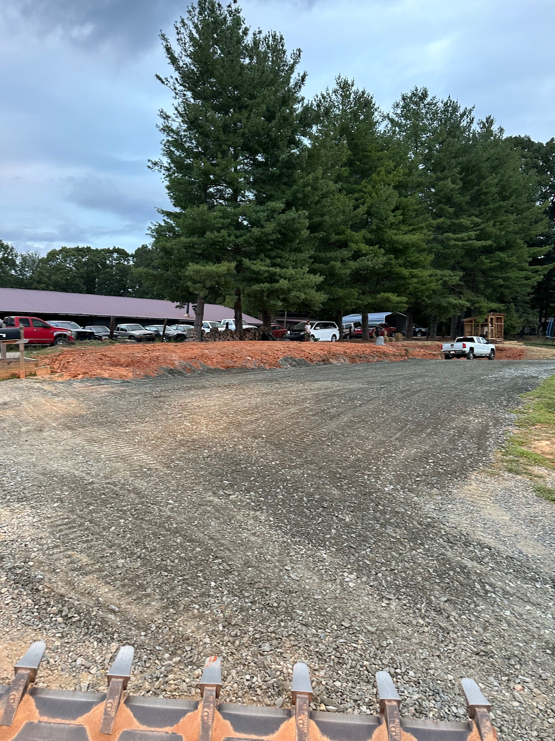 A view from construction equipment looking over a gravel lot toward a large shed with parked trucks and tall trees.