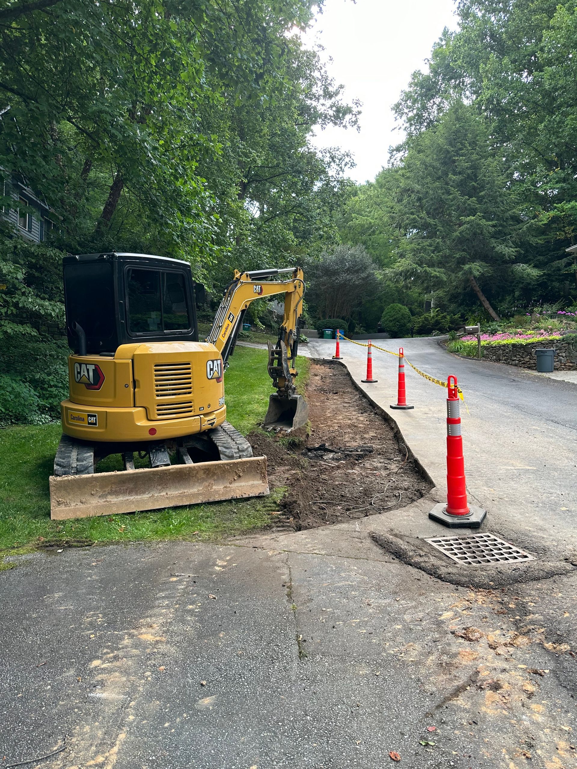 A yellow CAT mini excavator parked on a grassy verge next to a narrow road under construction, marked by orange cones.