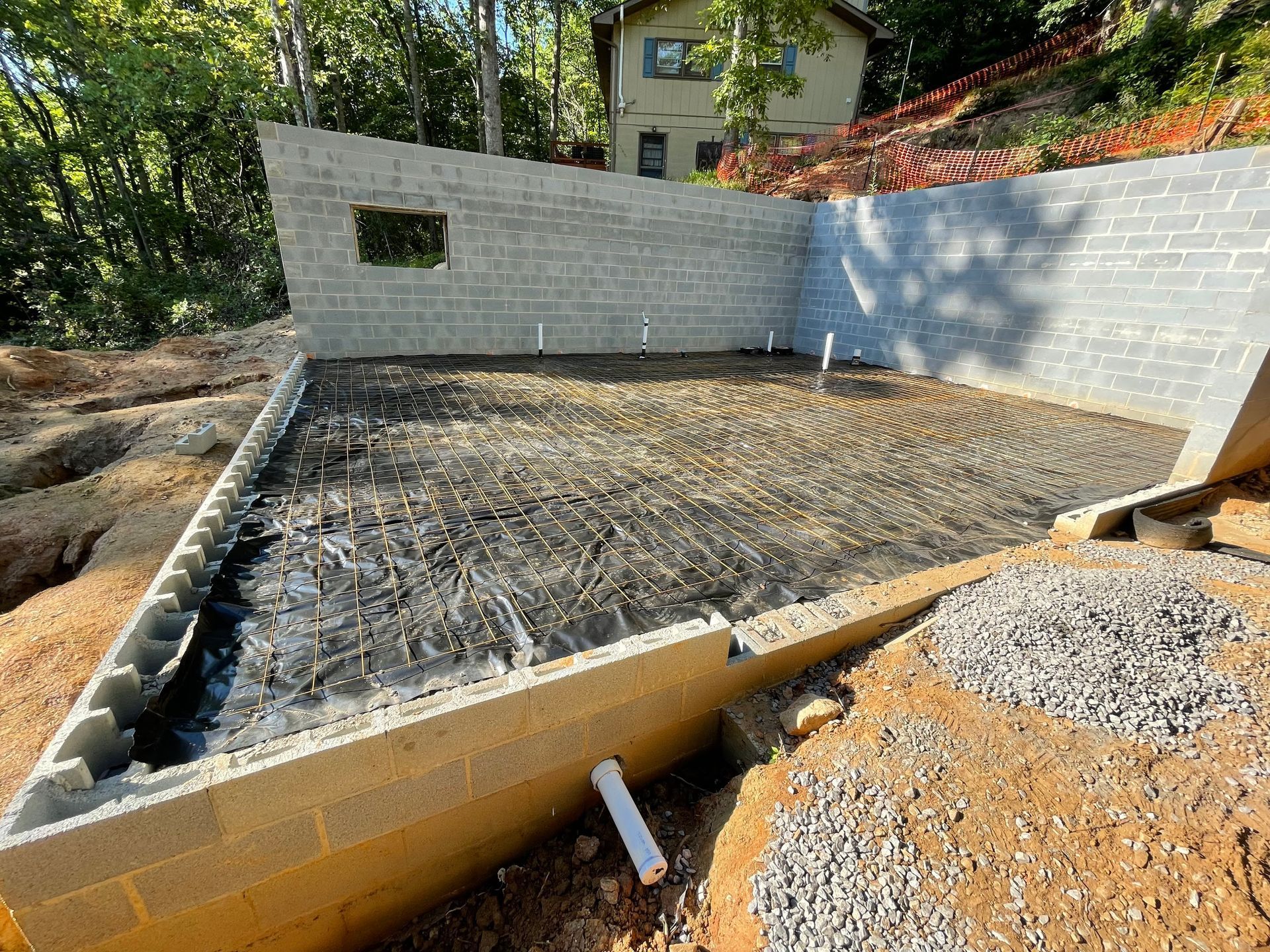Concrete foundation under construction with block walls, a gravel-covered floor, and a drainage pipe in a sloped yard.