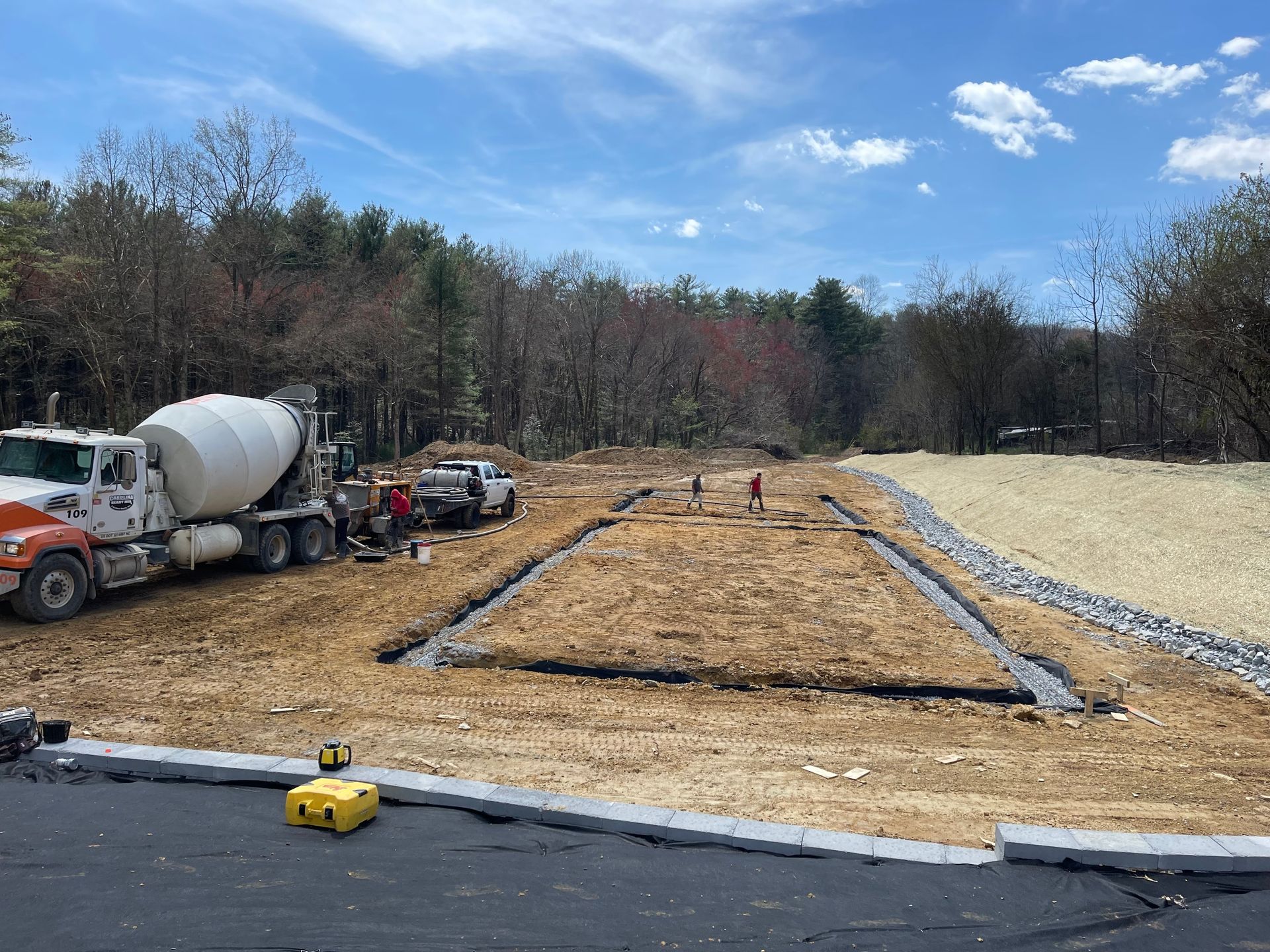 A construction site shows a cement truck parked by a rectangular trench foundation in a dirt clearing on a sunny day.