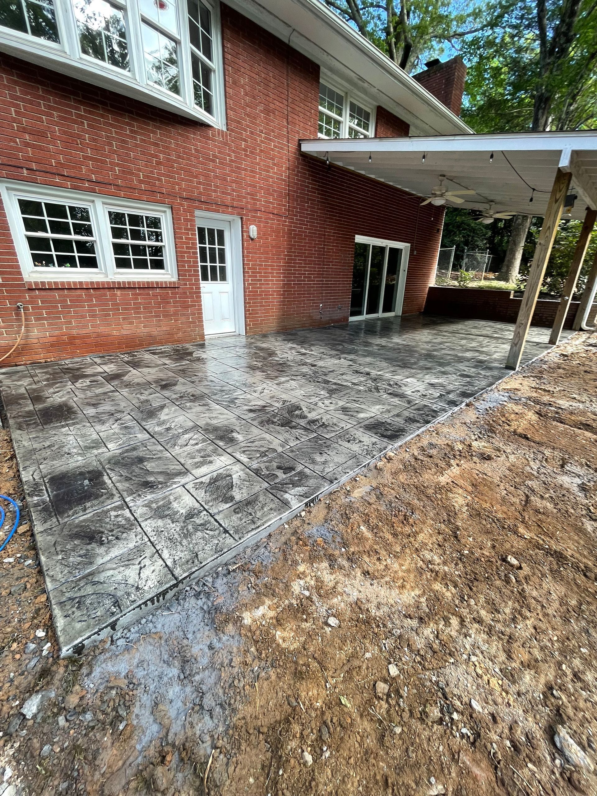 A red brick house with a new gray stamped concrete patio and a partially built wooden roof structure in the backyard.