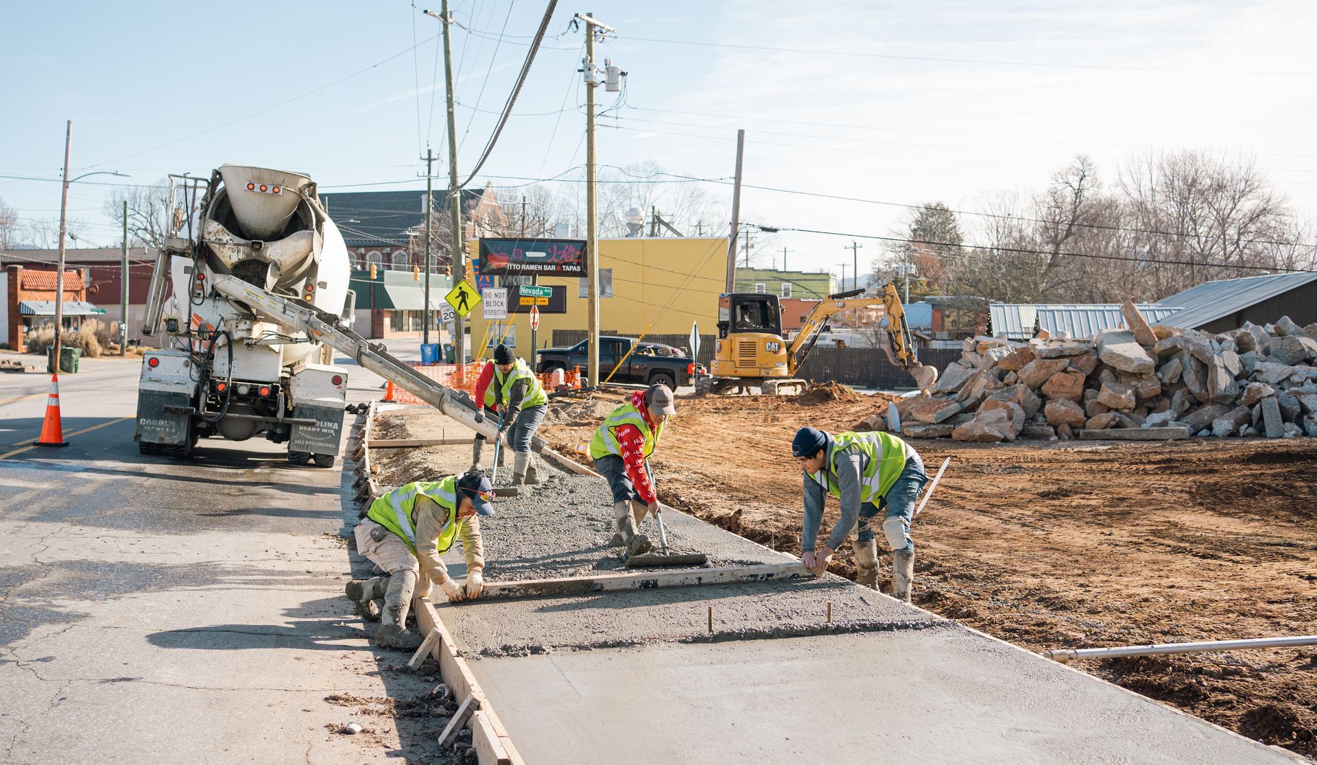 Construction workers in safety vests pour and smooth wet concrete for a new sidewalk alongside a cement truck.