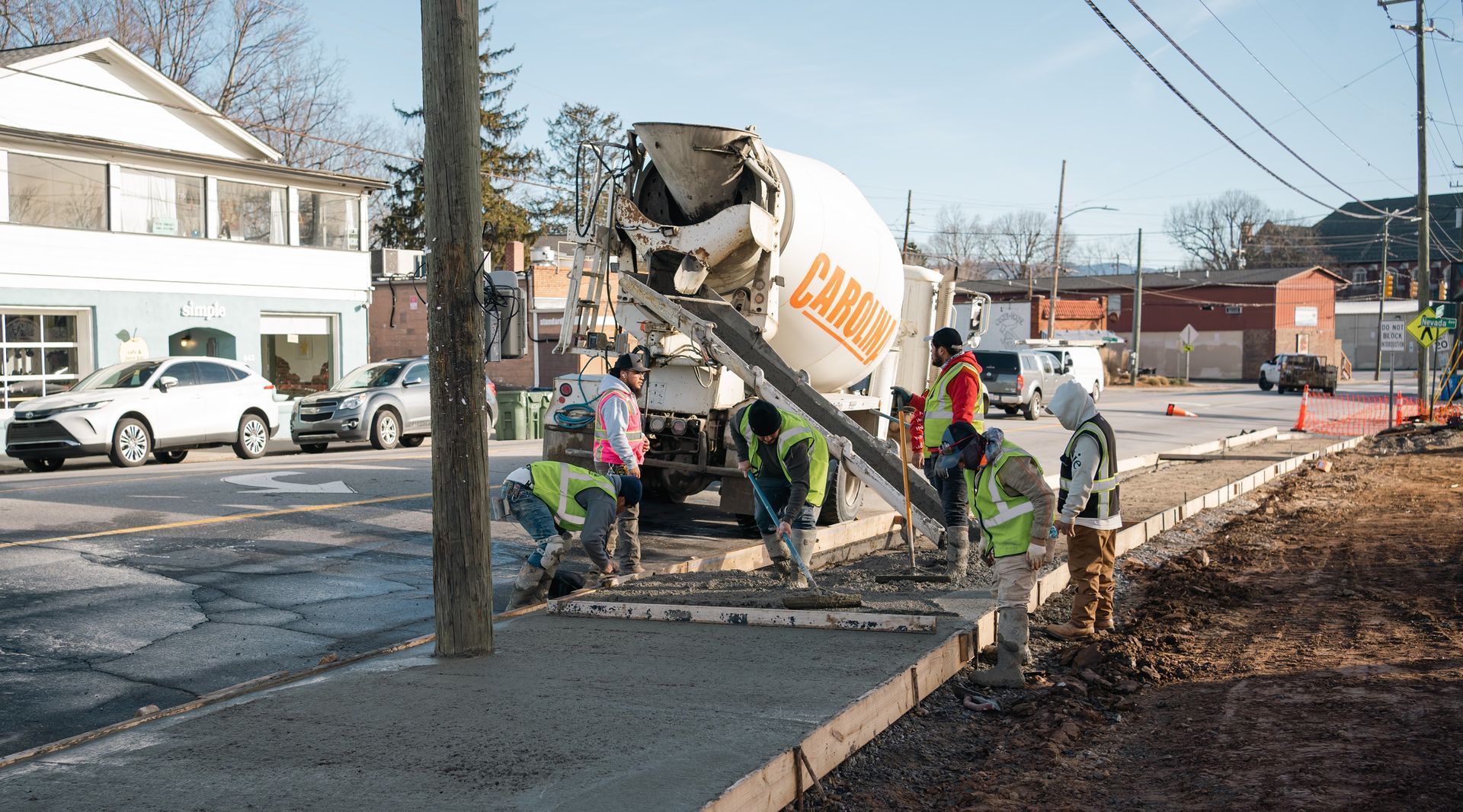 Construction workers pour wet concrete from a mixer truck to form a new sidewalk along a street.