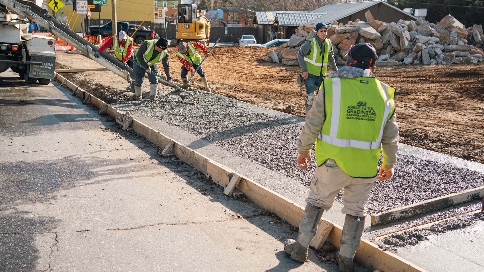 Construction workers pour and smooth concrete for a new sidewalk along a road, wearing high-visibility safety vests.