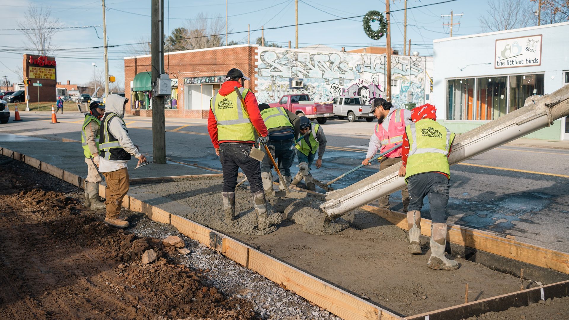 Construction workers in high-visibility vests pour and smooth concrete into a wooden frame on a street.