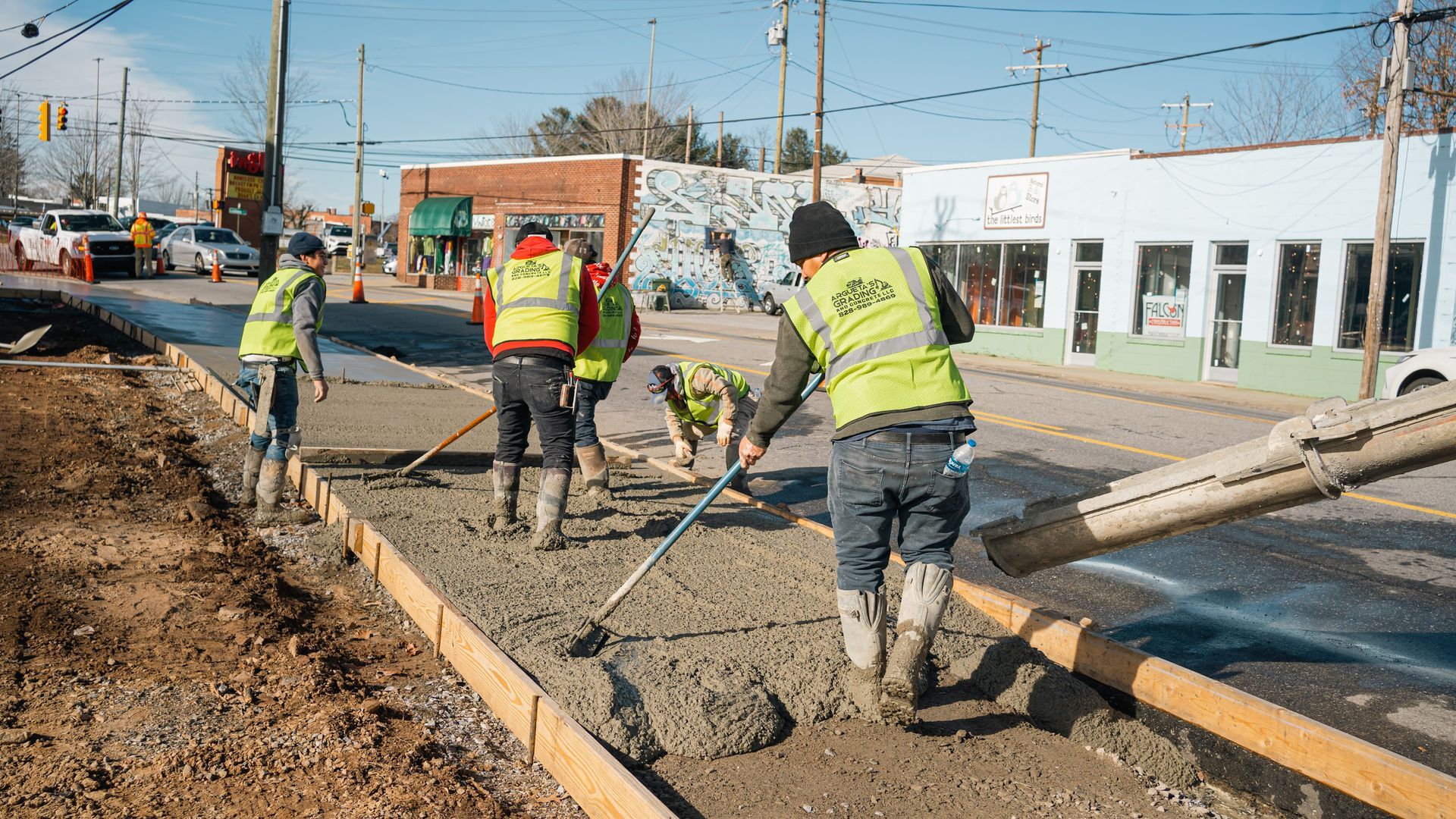 Construction workers in high-visibility vests pour and smooth wet concrete for a new sidewalk along a town street.