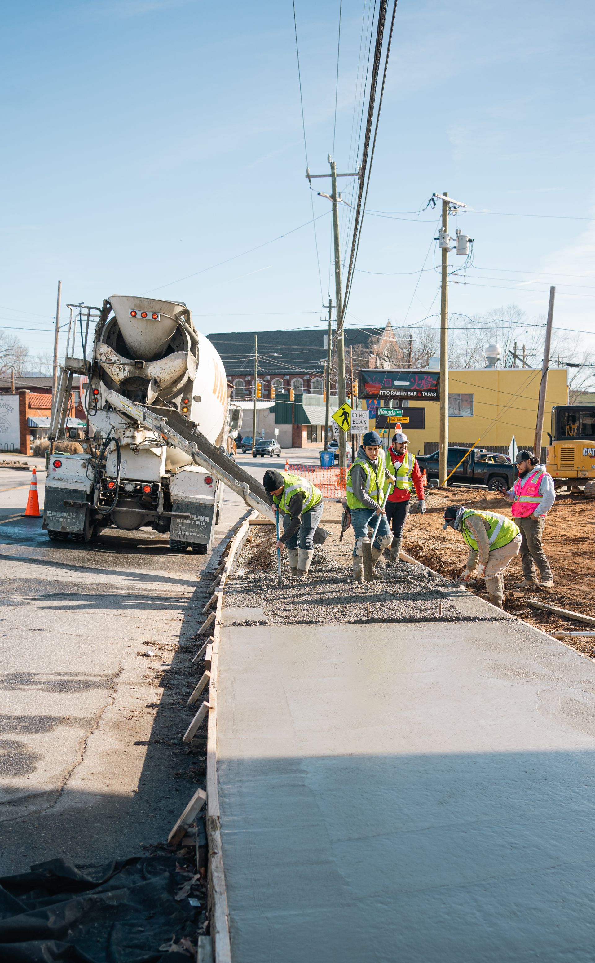 Construction workers in high-visibility vests pour wet concrete from a mixer truck to form a new sidewalk.