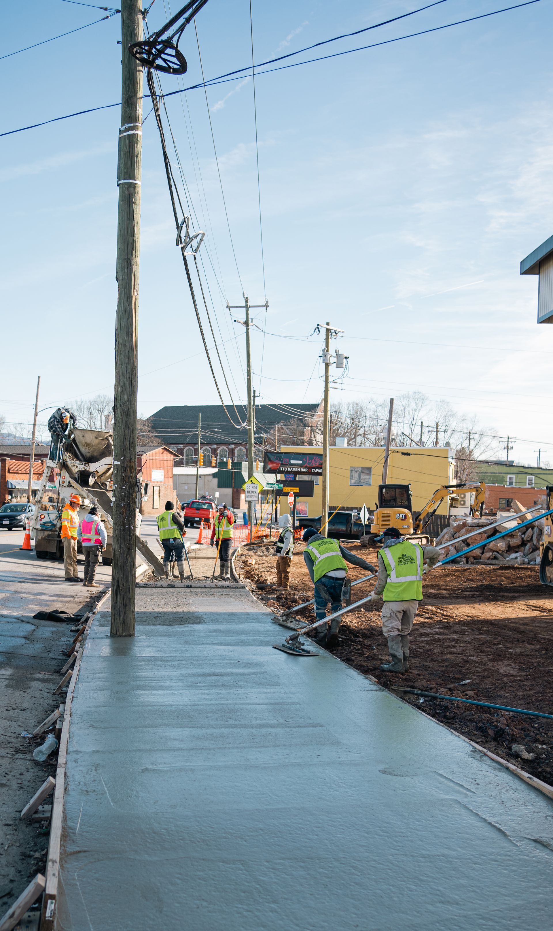 Construction workers in high-visibility vests finish pouring a new concrete sidewalk along a street.