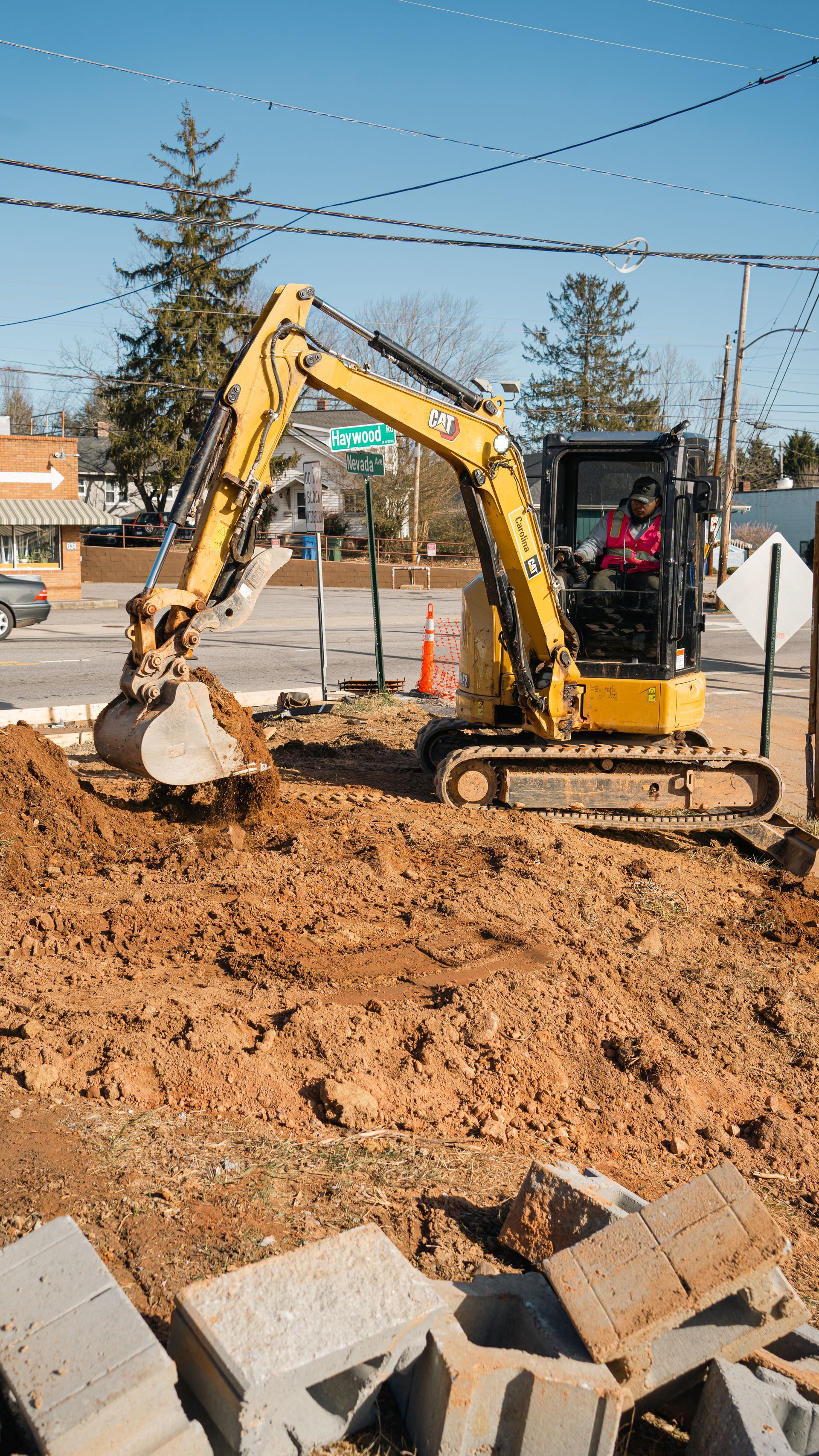 A yellow excavator digs into a patch of brown dirt at a construction site, with concrete blocks in the foreground.