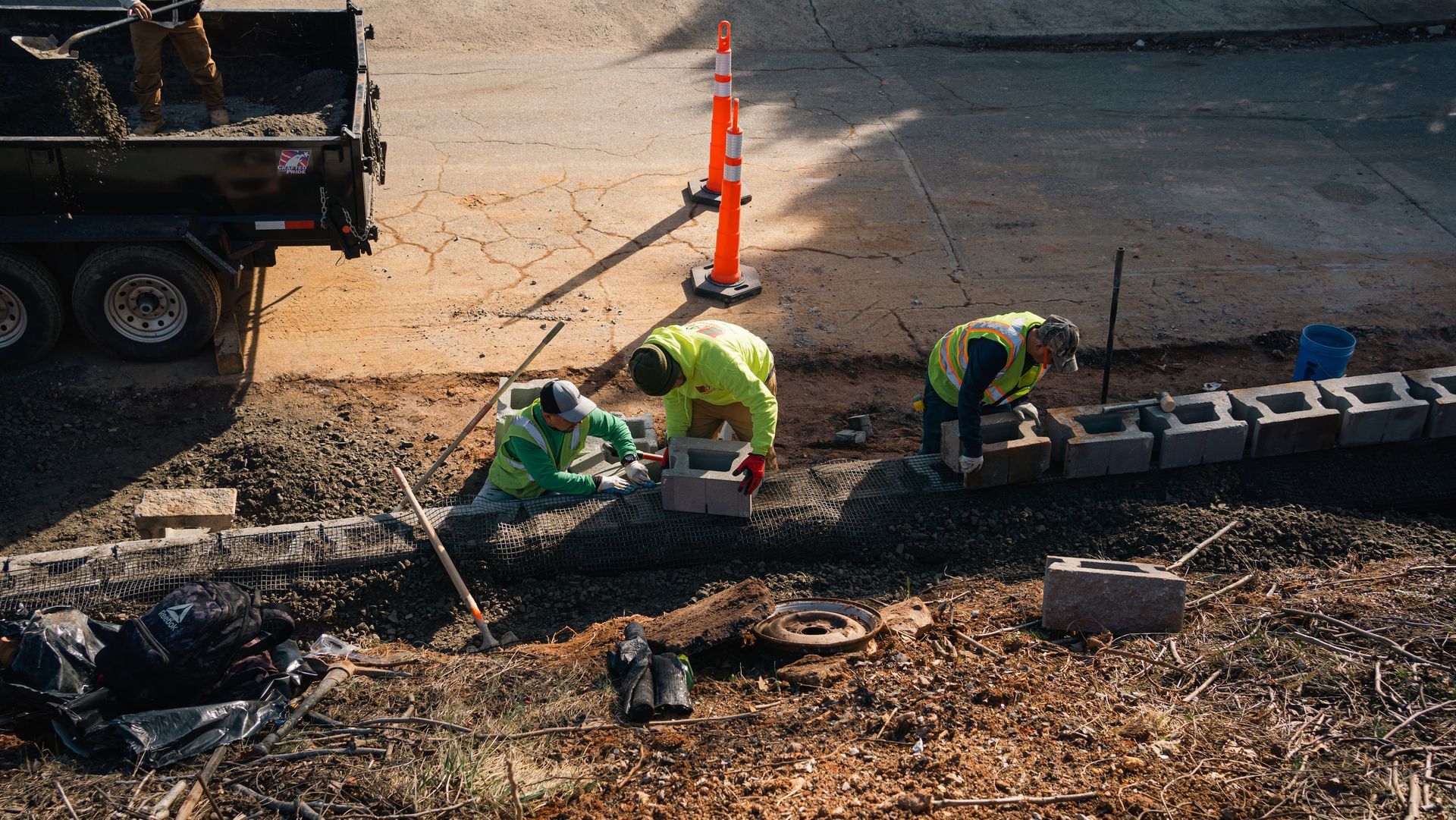 Construction workers in high-visibility vests install a concrete block retaining wall alongside a parked utility trailer.