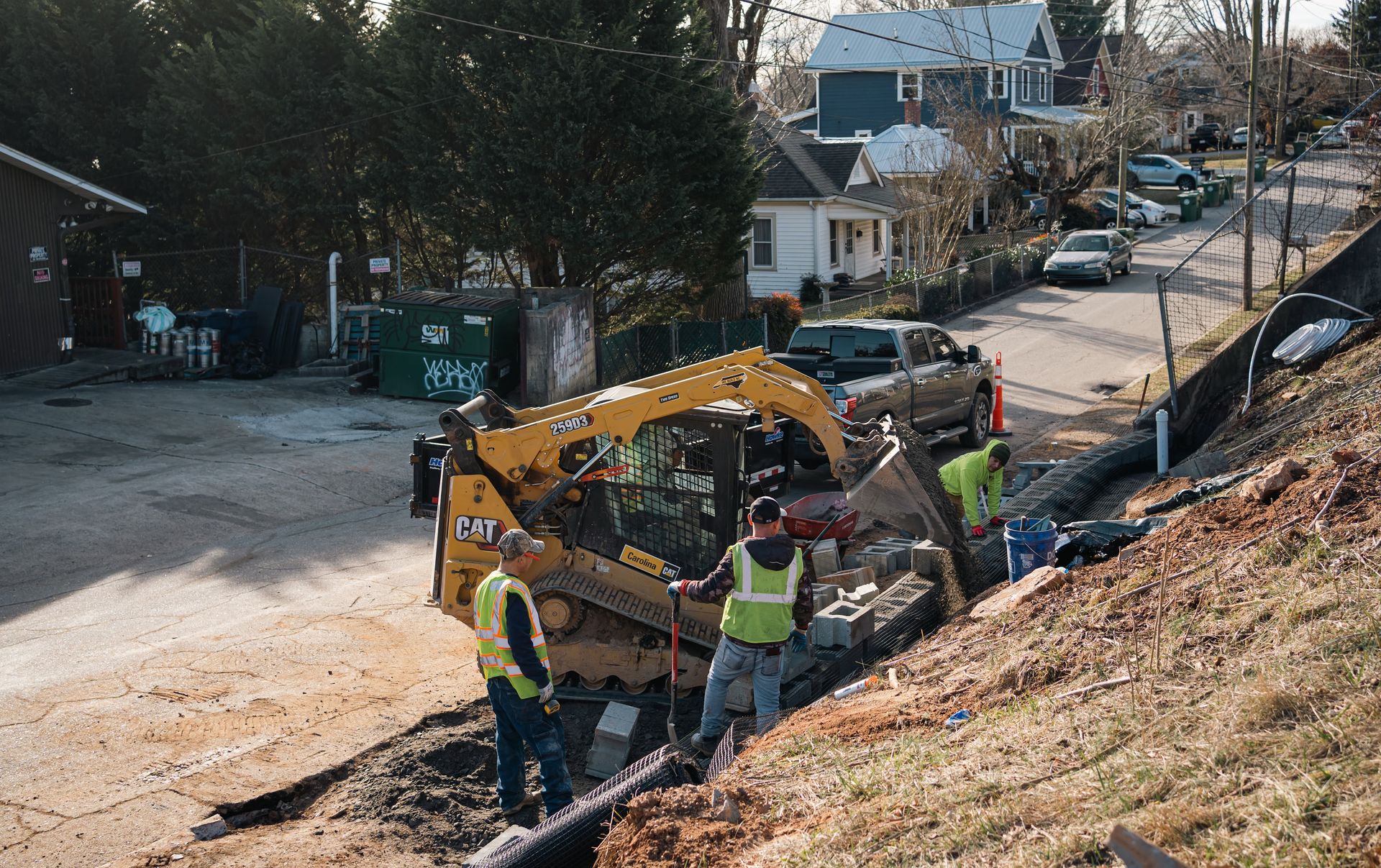Three construction workers operate a yellow CAT skid steer to install drainage piping along a residential roadside.