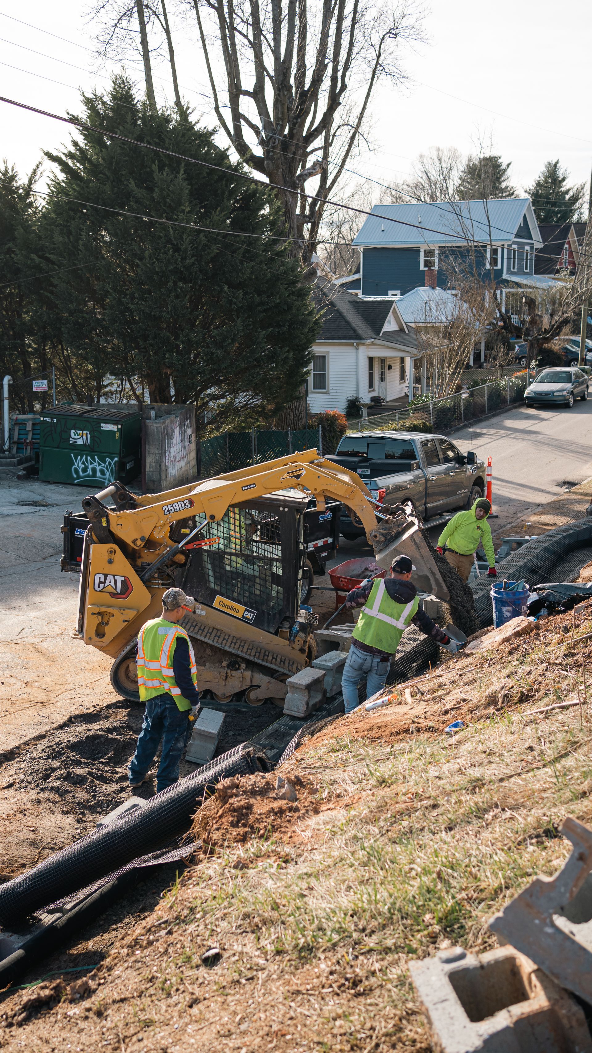 Workers in high-visibility vests use a yellow skid-steer loader to install concrete blocks on a residential roadside.