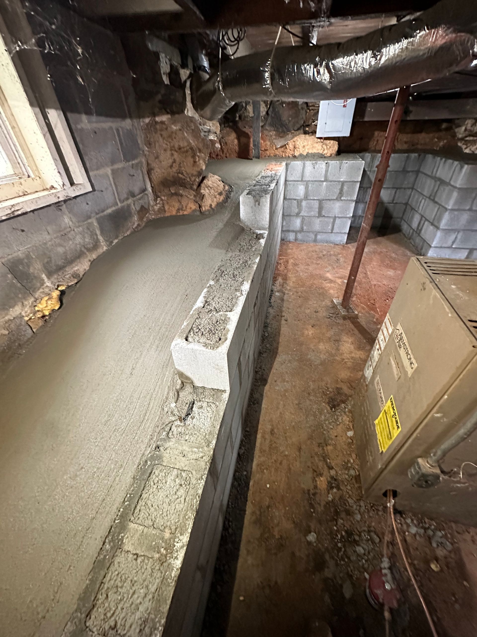 Fresh concrete footer poured along a basement wall beside a furnace and dirt floor in an unfinished crawl space.