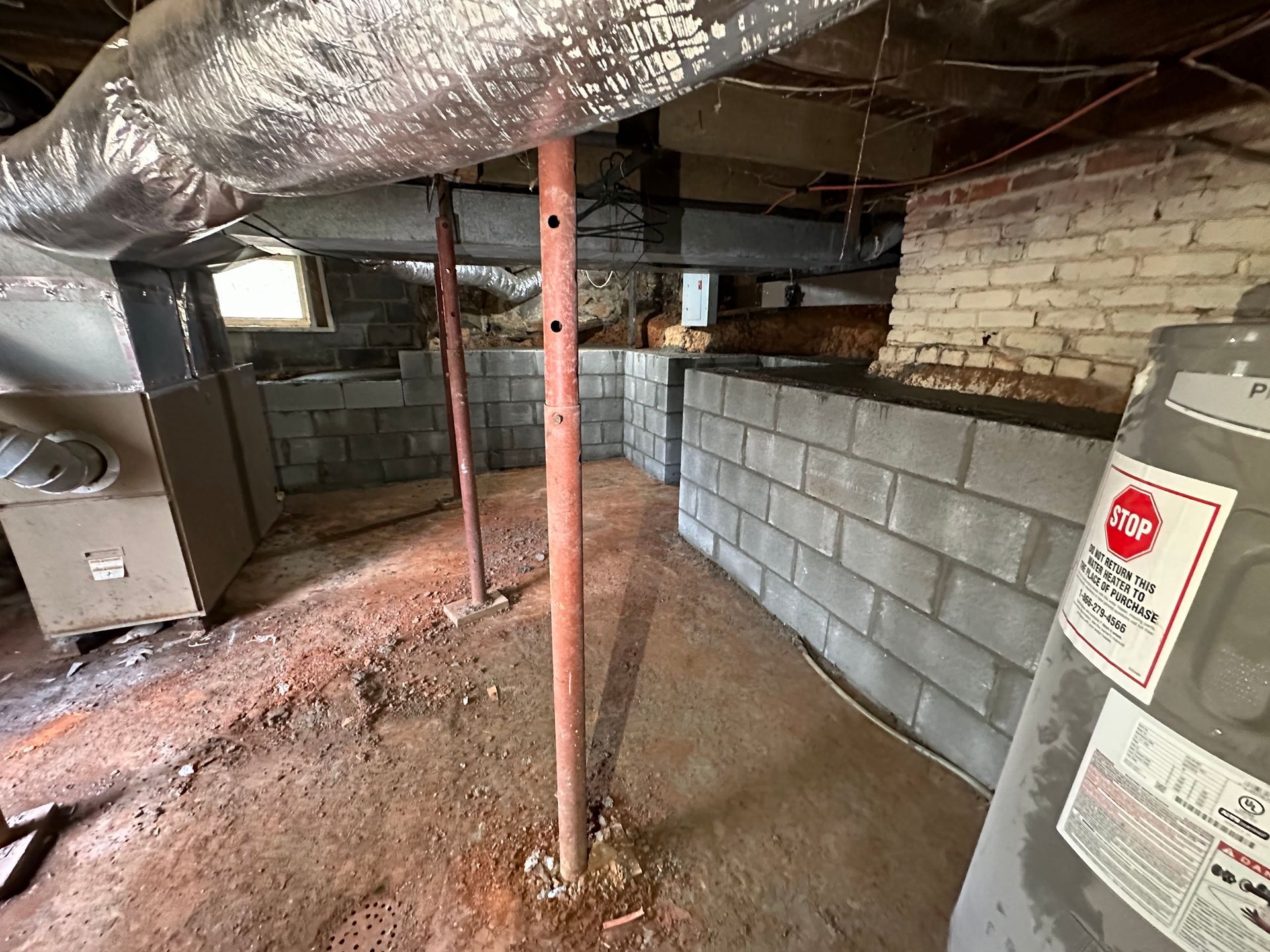 A crawlspace with a dirt floor, metal support columns, a gray block foundation wall, HVAC ductwork, and a water heater.