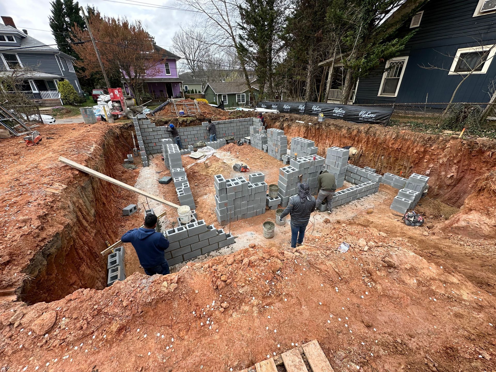 Workers build concrete block foundation walls in a large, deep construction excavation surrounded by residential homes.