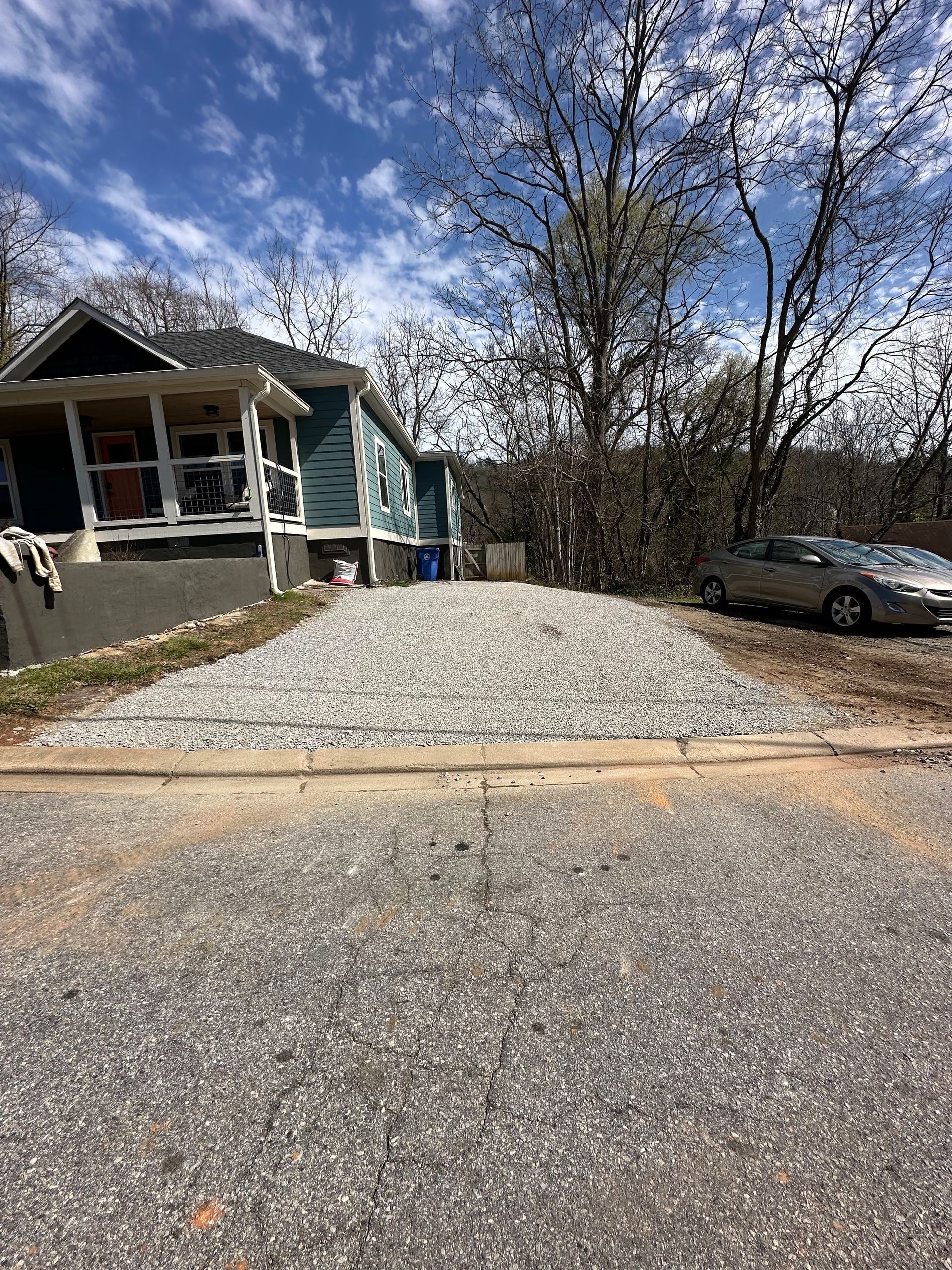 A gravel driveway leading up to a blue house with a porch, seen from the street on a sunny day with trees in the background.