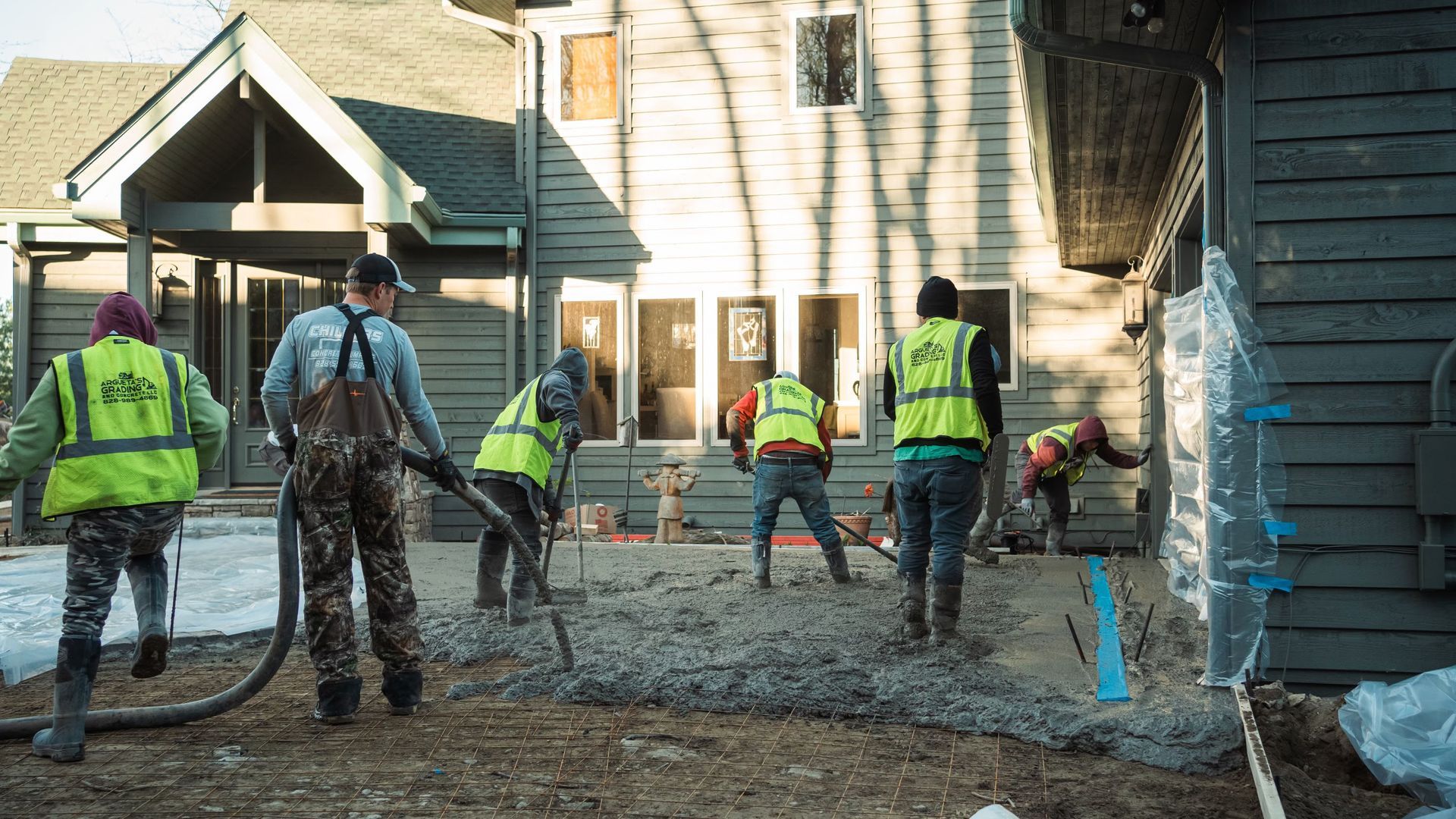 Construction workers in high-visibility vests work on a concrete foundation in front of a residential house.