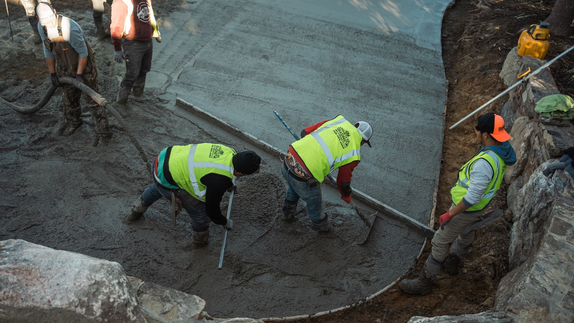 Construction workers wearing high-visibility vests use tools to smooth wet concrete at an outdoor site.