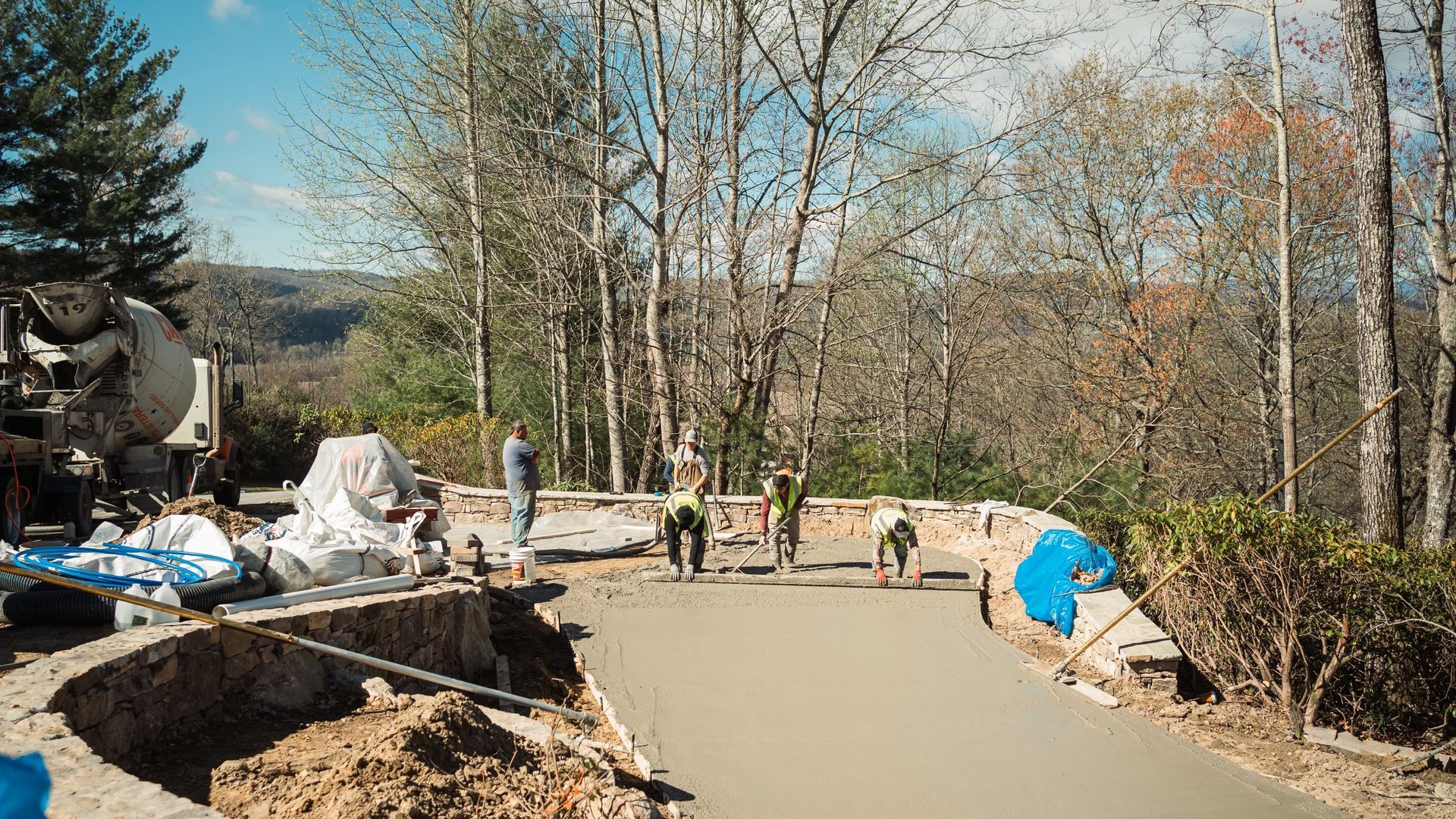 Workers smooth wet concrete for a new driveway at a hillside construction site with a cement truck parked nearby.