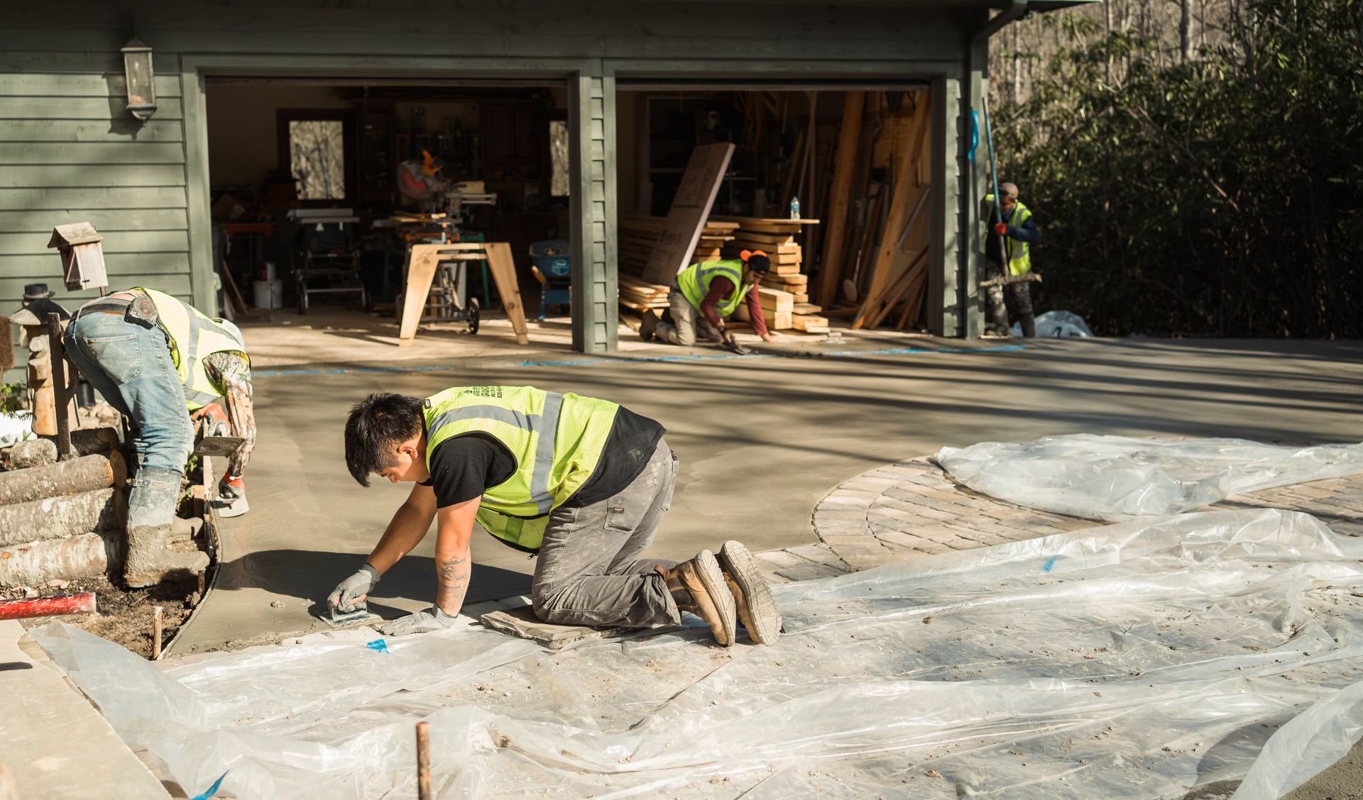 Construction workers wearing high-visibility vests finish a concrete driveway in front of an open garage.