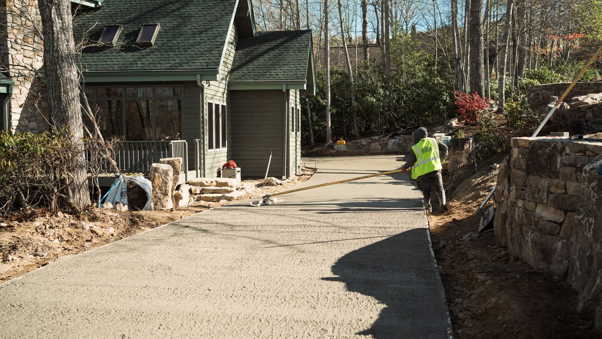 A person in a high-visibility vest stands on a newly poured concrete driveway near a house in a wooded setting.