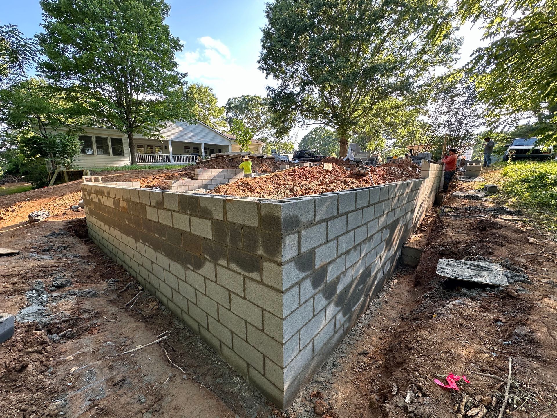 A rectangular concrete block foundation wall stands on a construction site surrounded by trees and dirt.