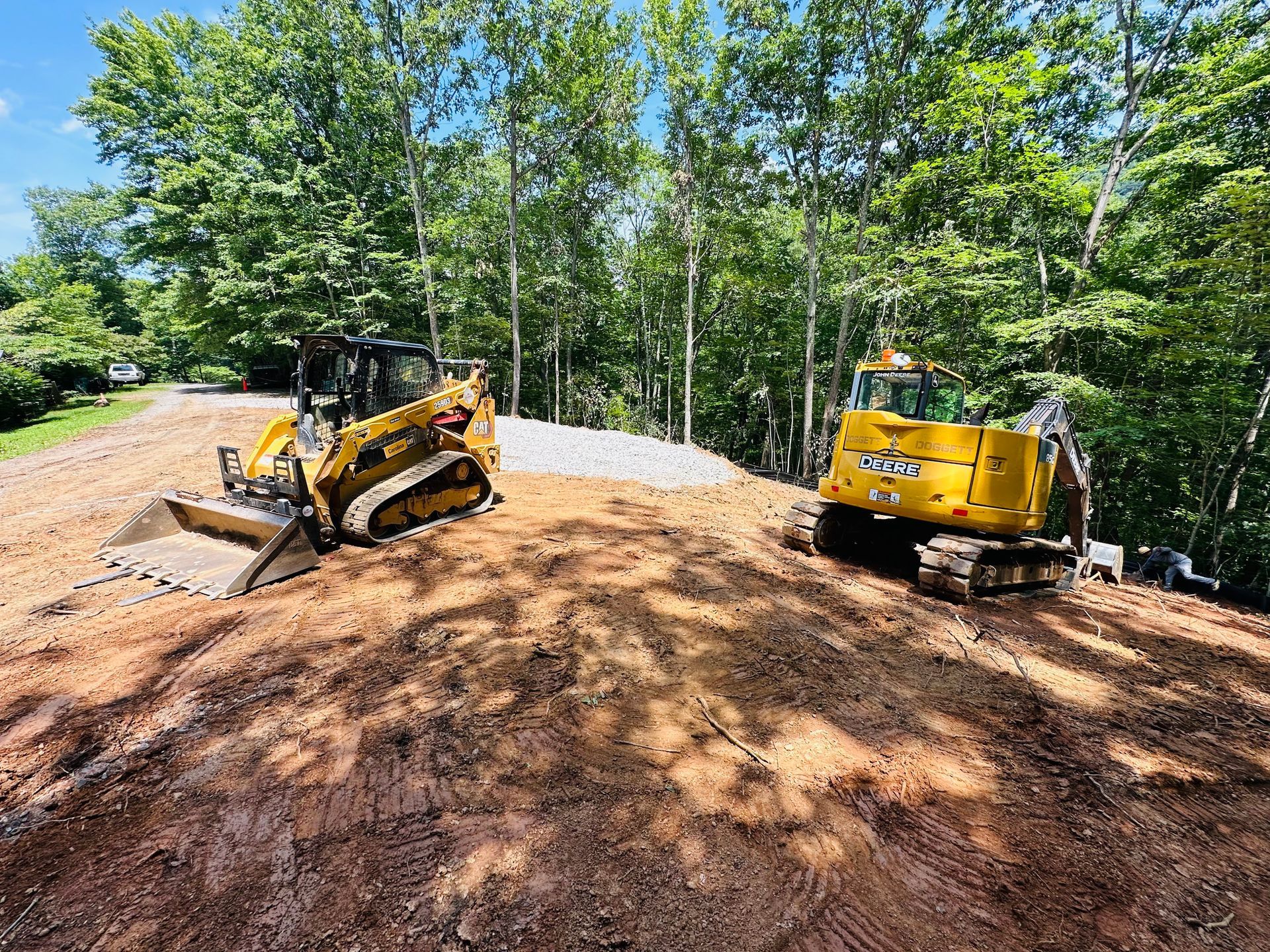 A yellow skid steer and a yellow excavator sit on a dirt construction site surrounded by trees on a sunny day.