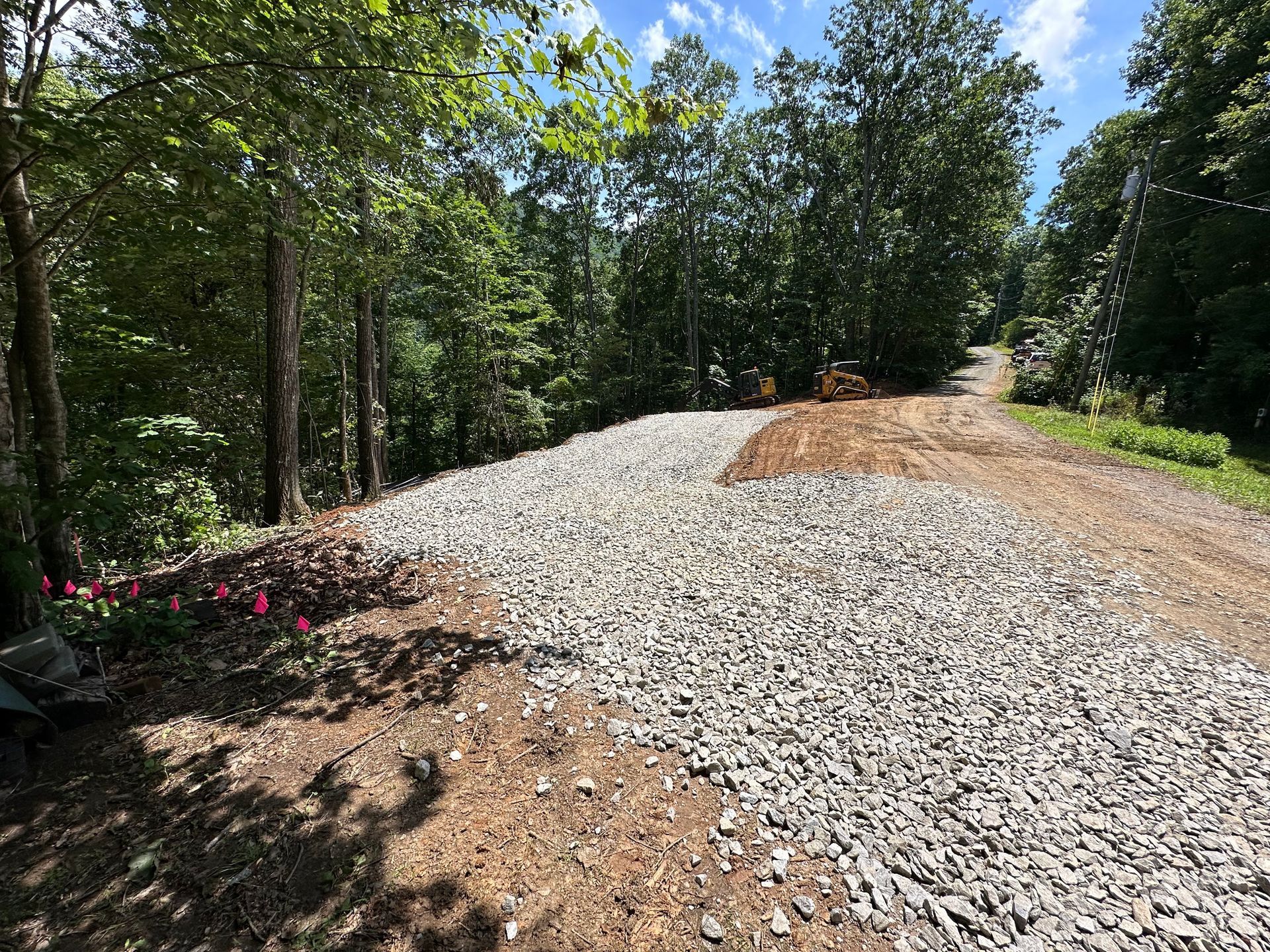 An excavator bucket in the foreground overlooks a gravel-covered work site with dirt piles, trees, and vehicles nearby.
