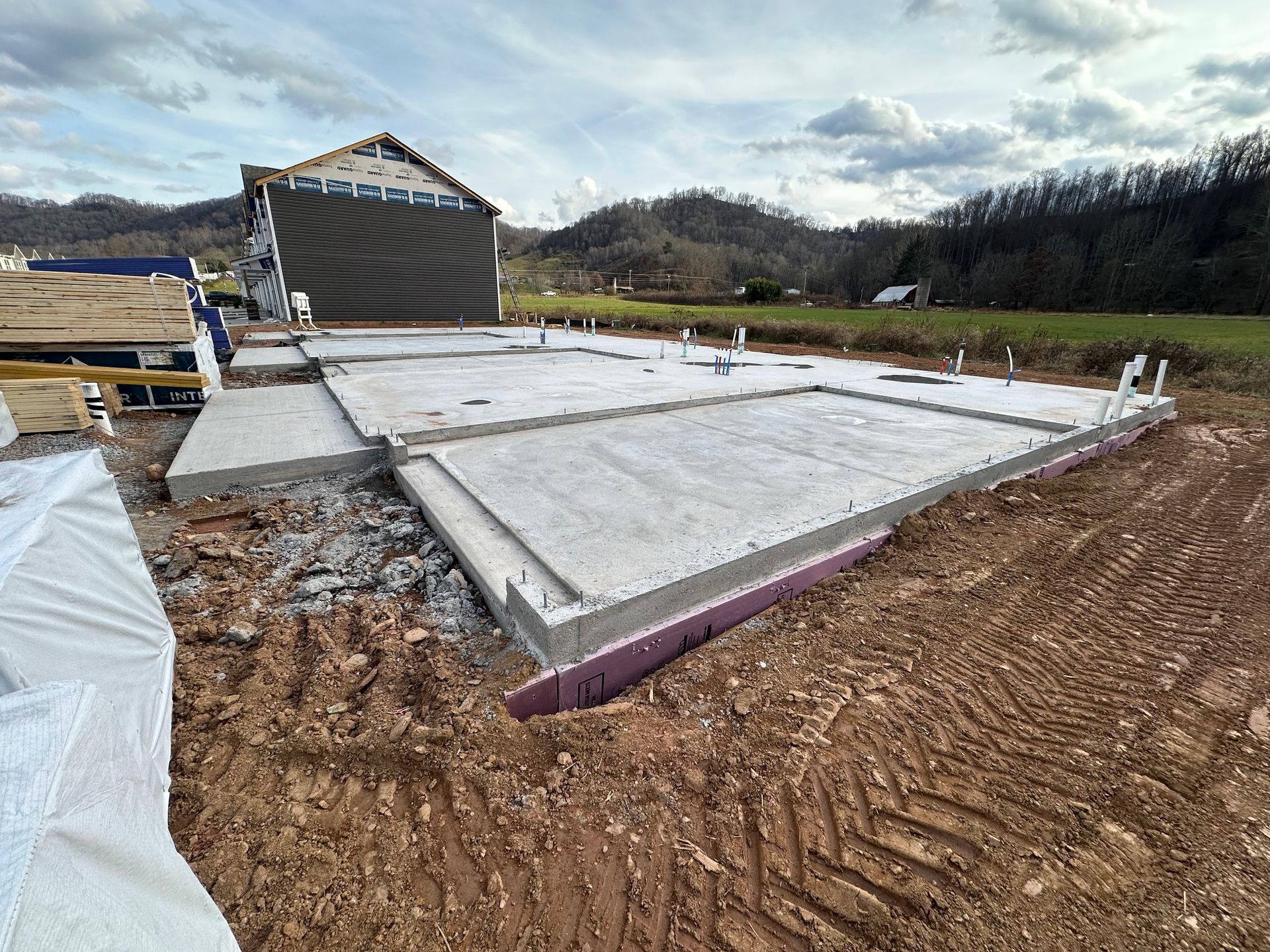 A concrete foundation for a house under construction in a rural, grassy field under a cloudy sky.