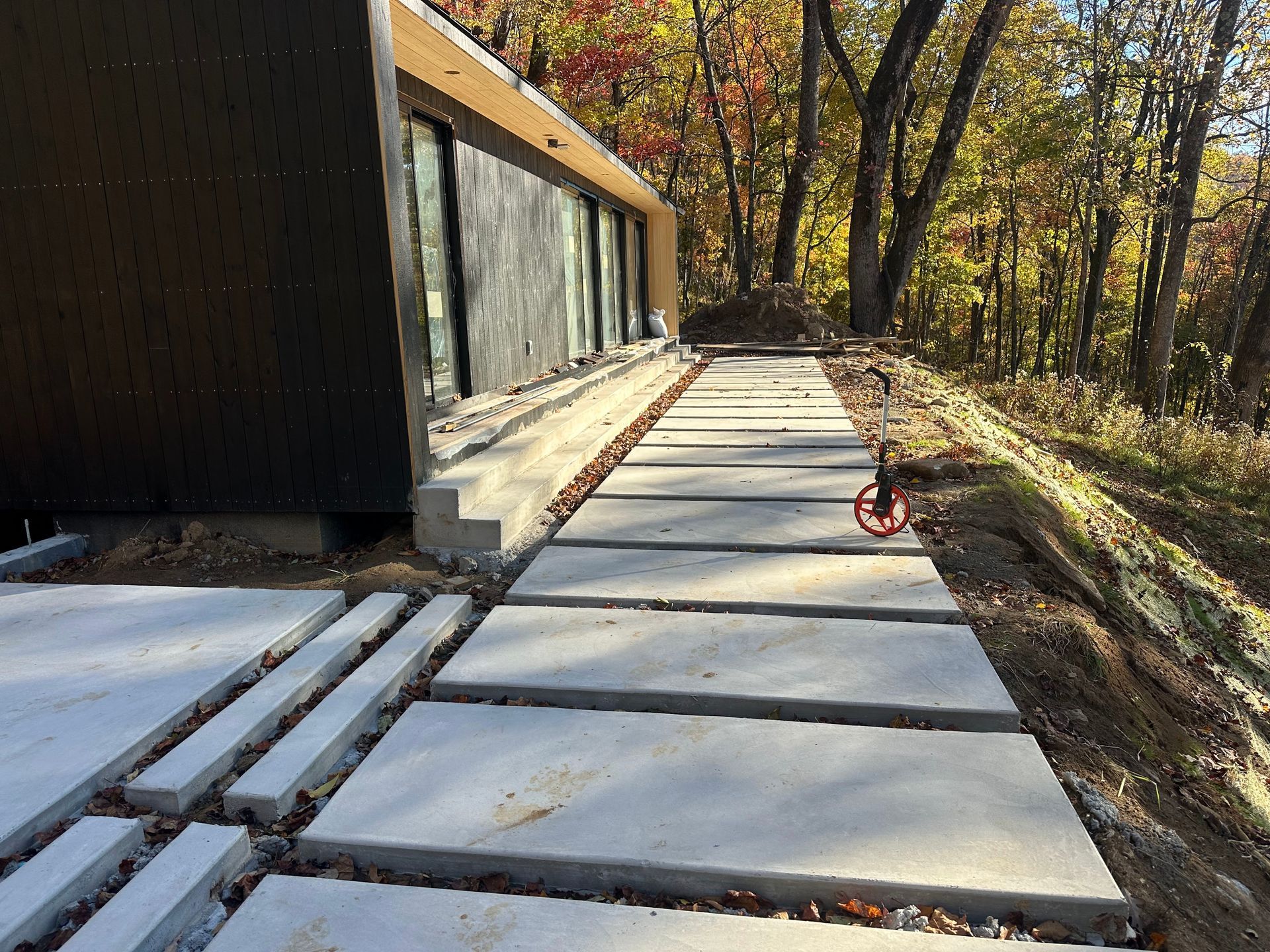 A long concrete paver walkway runs alongside a modern black building with glass doors, bordering a wooded area in autumn.