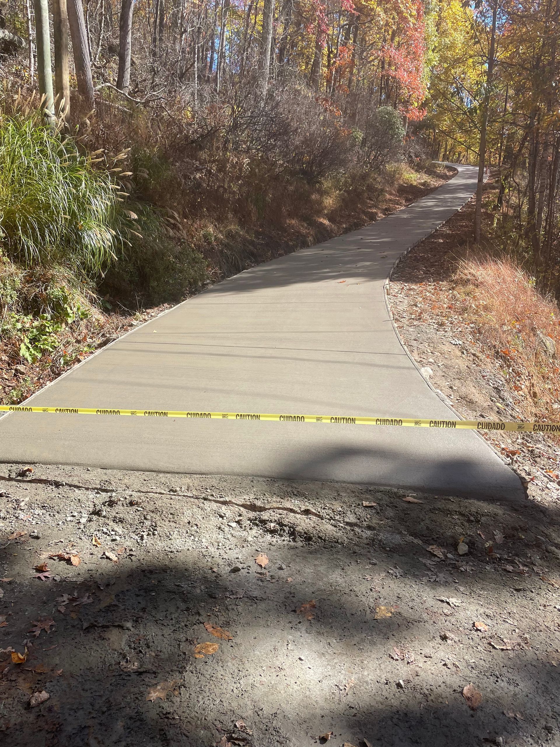A newly paved concrete path winding through an autumn forest, blocked off by yellow caution tape.