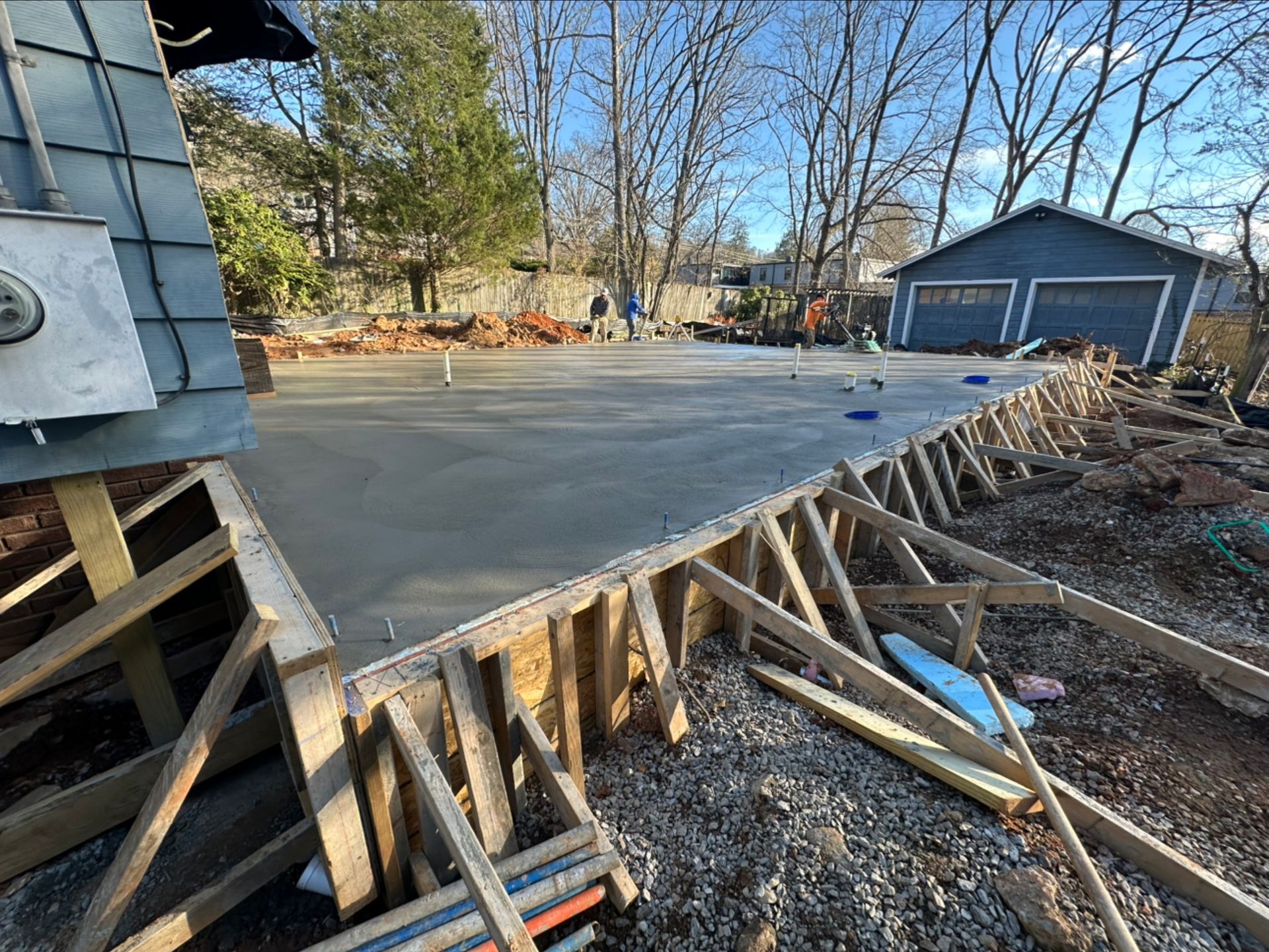 Freshly poured concrete slab foundation with wooden formwork in a backyard setting under a clear blue sky.