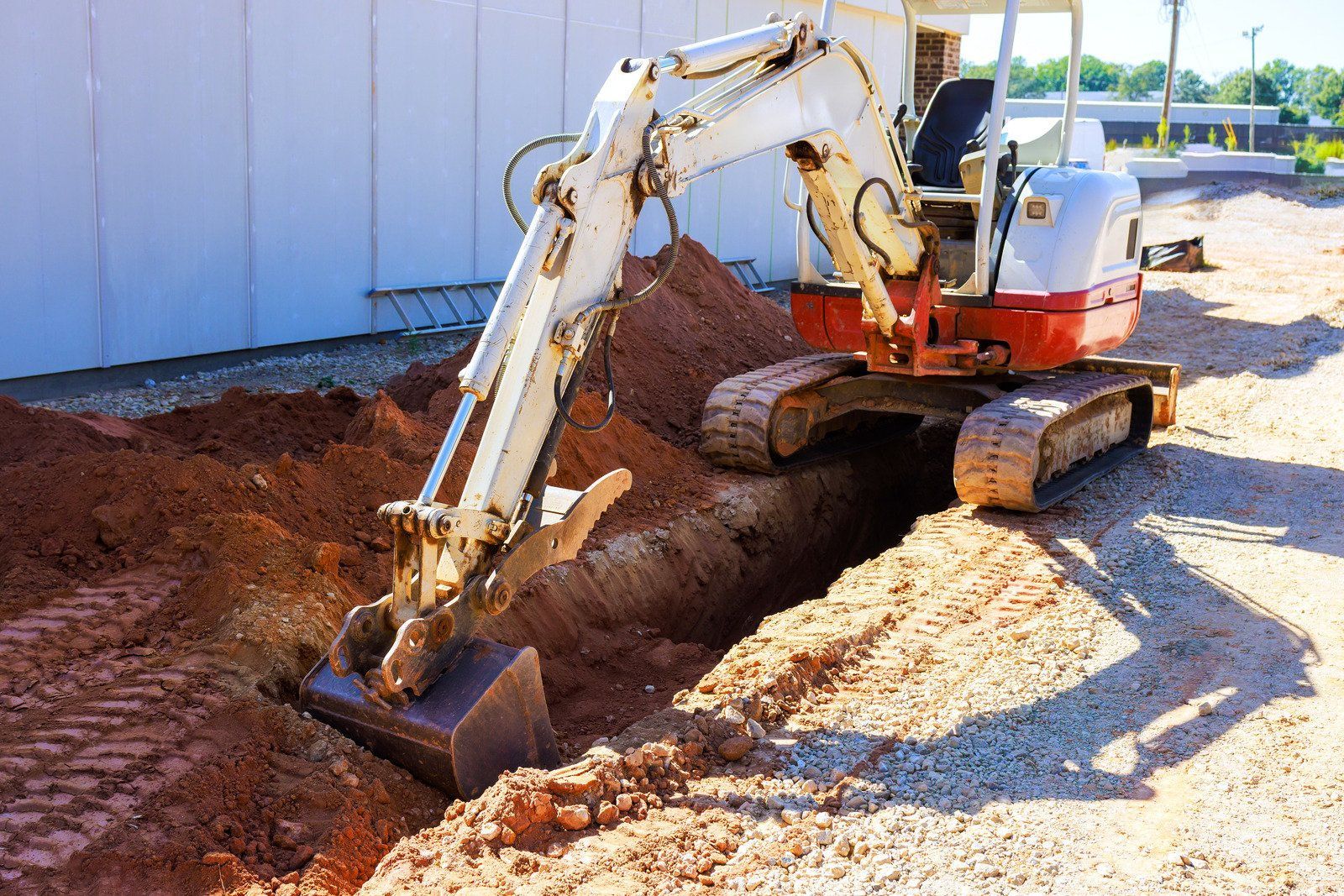 A small white and orange excavator digging a trench in red dirt next to a building wall.