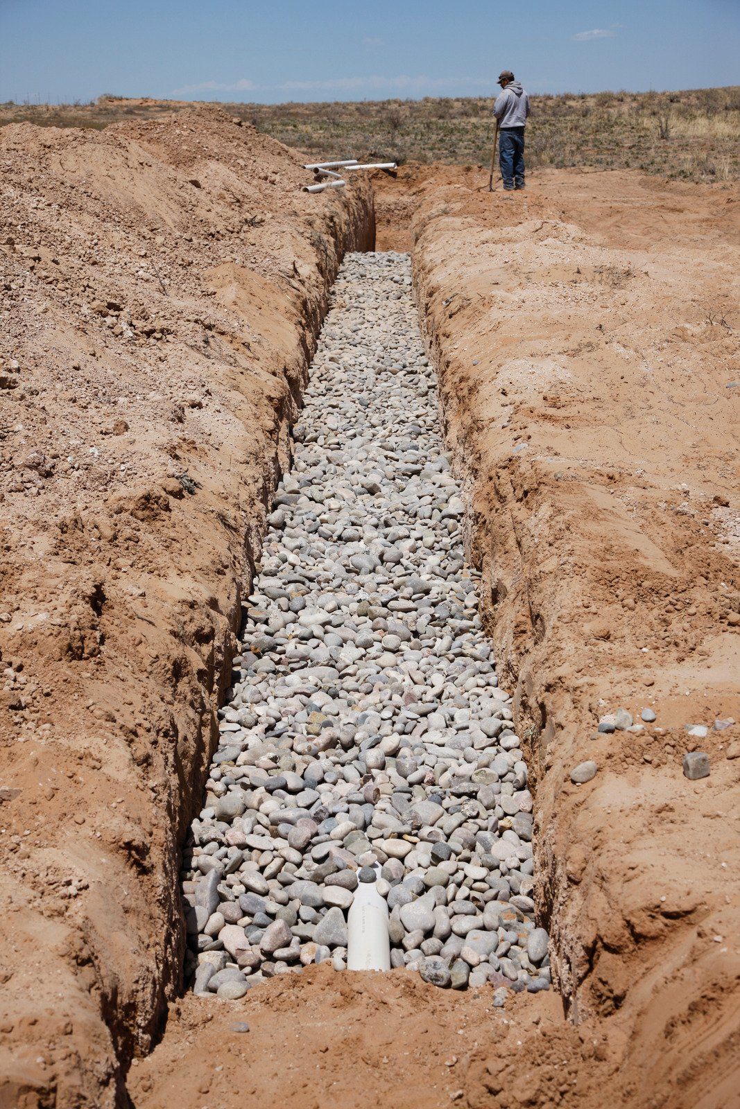 A person stands near a deep trench in dry, reddish soil filled with grey gravel and a white drainage pipe.