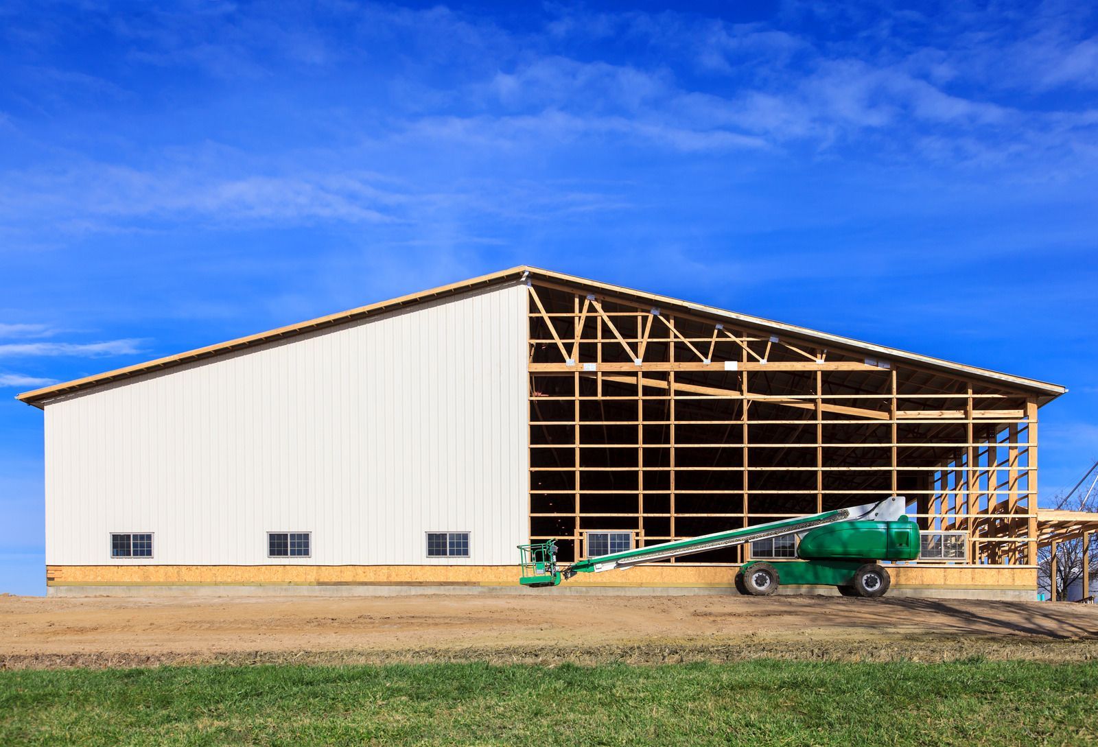 A large, partially sided metal building under construction with a green boom lift parked in front against a blue sky.