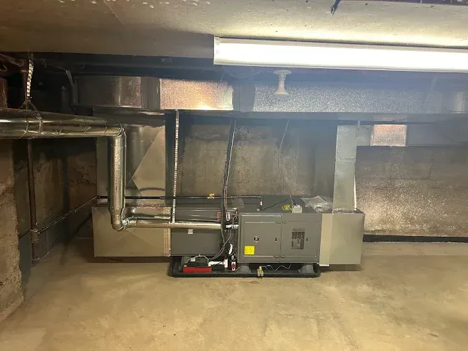 Furnace and ductwork in a basement. Grey metal components, concrete floor, overhead fluorescent light.
