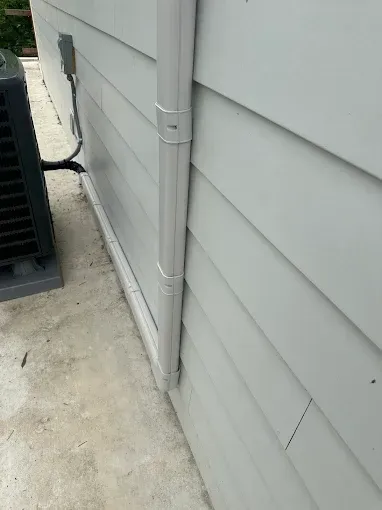 Gray gutter and downspout on a light gray sided building next to an AC unit.