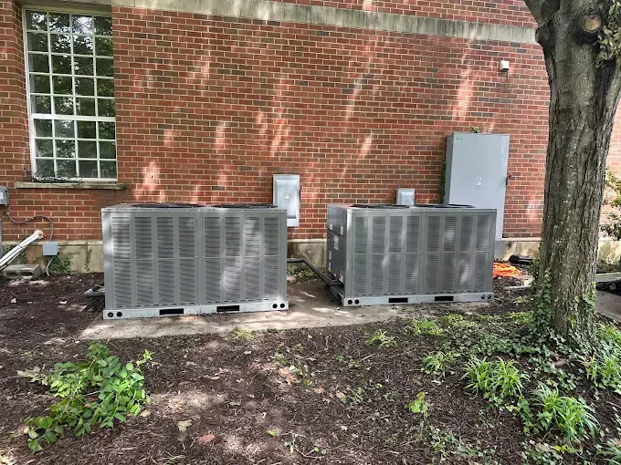 Two air conditioning units sit in front of a brick building near a tree.
