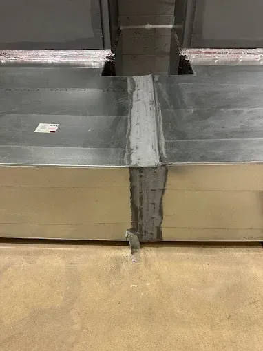 Metal ductwork joint sealed with sealant, resting on a concrete floor in a room.