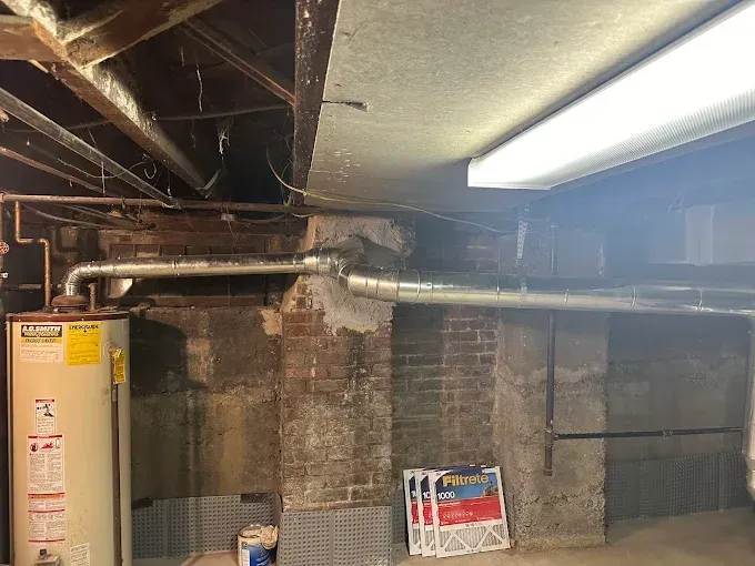 Basement interior with water heater, ductwork, exposed brick and concrete walls, and fluorescent lighting.