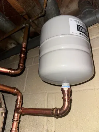 Copper pipes connected to a white expansion tank in a basement, mounted on a cinder block wall.