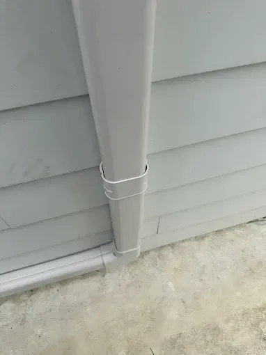 Gray downspout attached to a light gray siding, secured by a white metal strap.