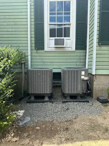 Two gray air conditioning units next to a green house, window above them.