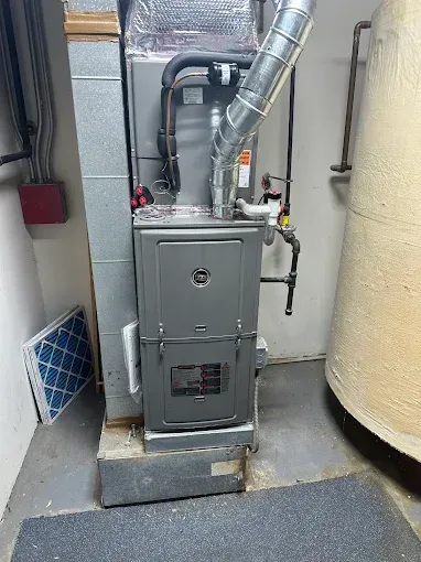 A furnace in a basement setting with ductwork, water heater, and filter. Gray metal.