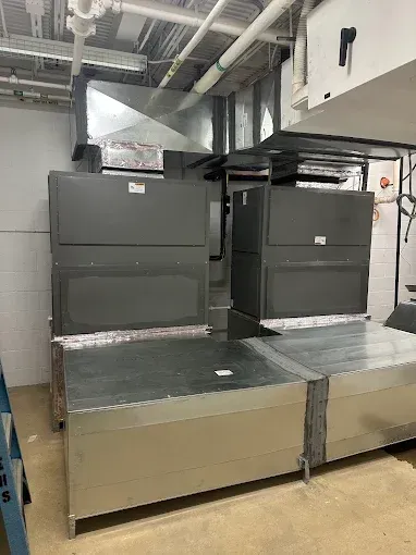 HVAC equipment in a mechanical room, featuring two large gray units connected to ductwork and ceiling pipes.