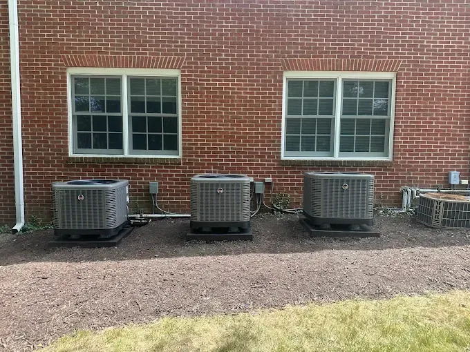 Three air conditioning units outside a brick building with two windows.
