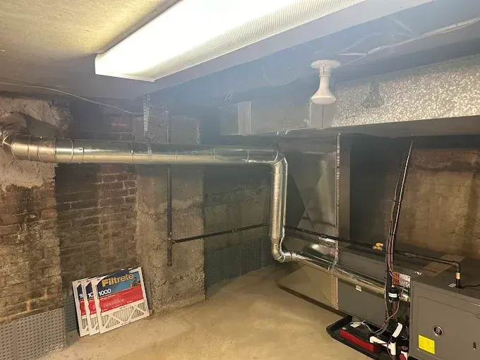 Basement with HVAC system, exposed ductwork, brick and concrete walls, fluorescent lighting.
